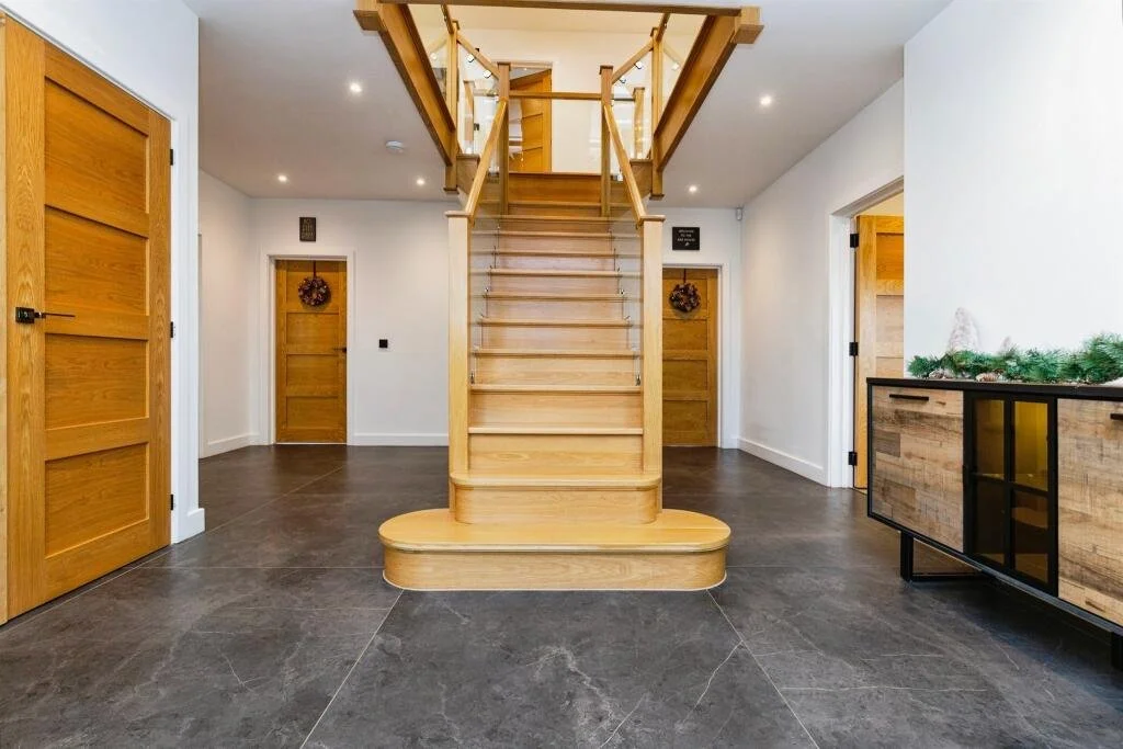 Interior view of a house with a wooden staircase leading to an upper floor, four wooden doors decorated with wreaths, dark tile flooring, and a wooden sideboard with a garland on top.