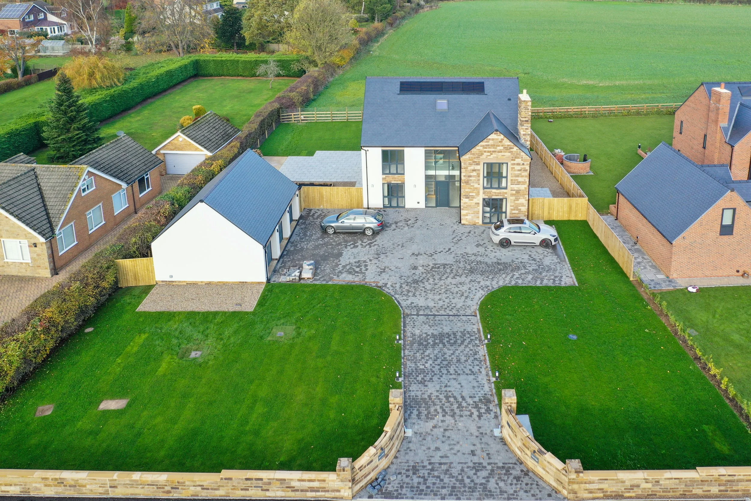 Aerial view of a modern residential property with a large stone and white house, a wide driveway, two parked cars, a large backyard with a lawn, and surrounding fences. Neighboring houses and green fields are visible.