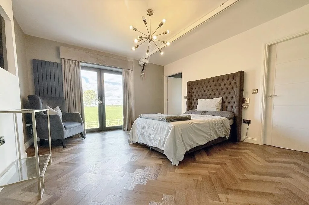 Bedroom with a large bed featuring a tufted headboard, a glass sliding door with curtains leading to an outdoor view, a gray armchair with a pillow, and a modern chandelier ceiling light fixture.