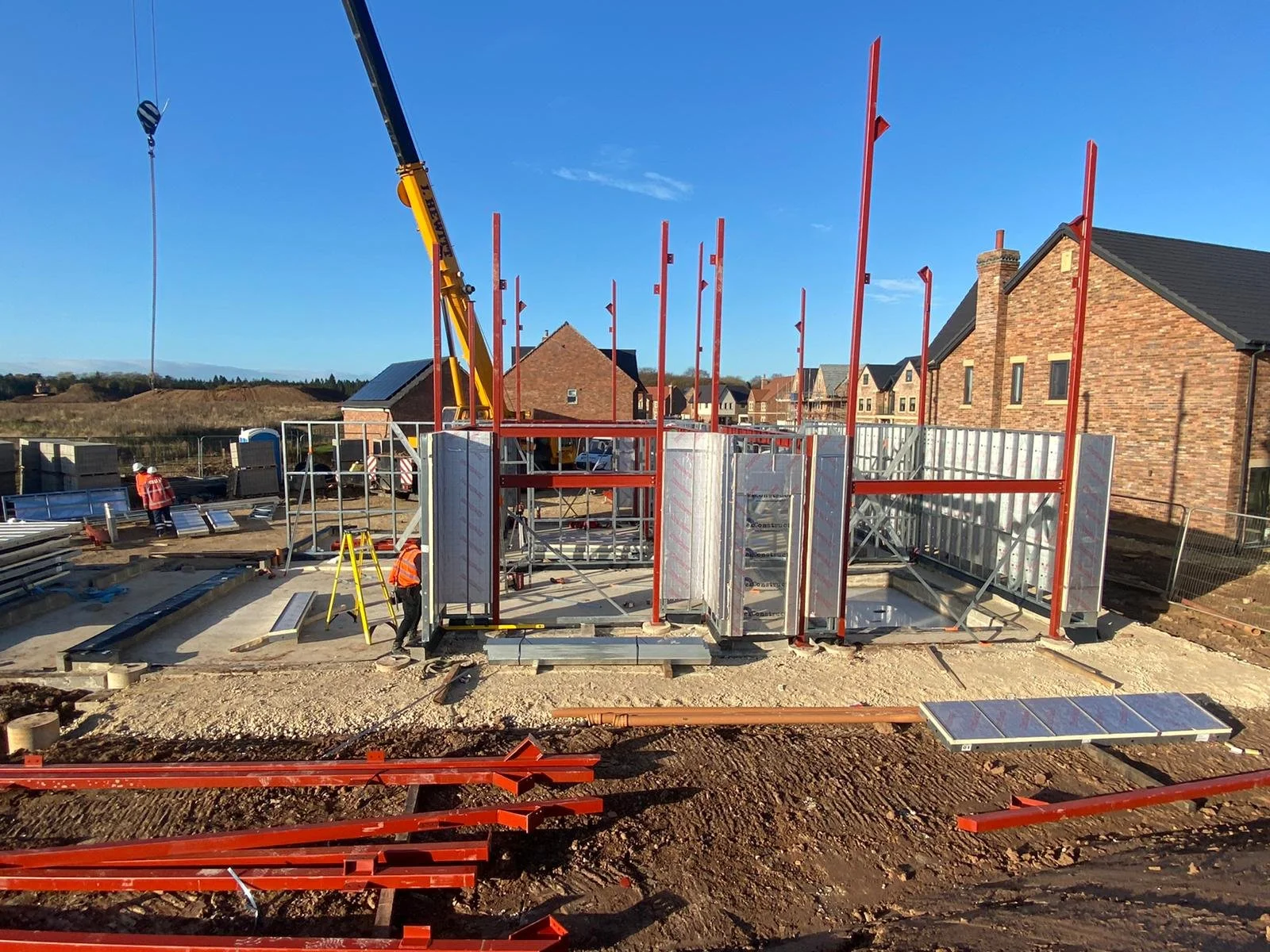 Construction site with scaffolding being assembled, workers wearing safety vests and helmets, and a crane. Houses in the background under a clear blue sky.