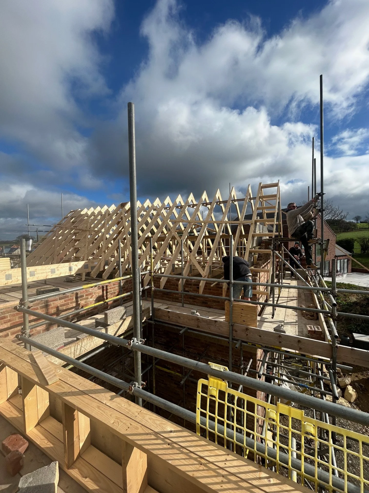 Construction site with workers building a house, visible wooden framework and scaffolding, against a partly cloudy sky.