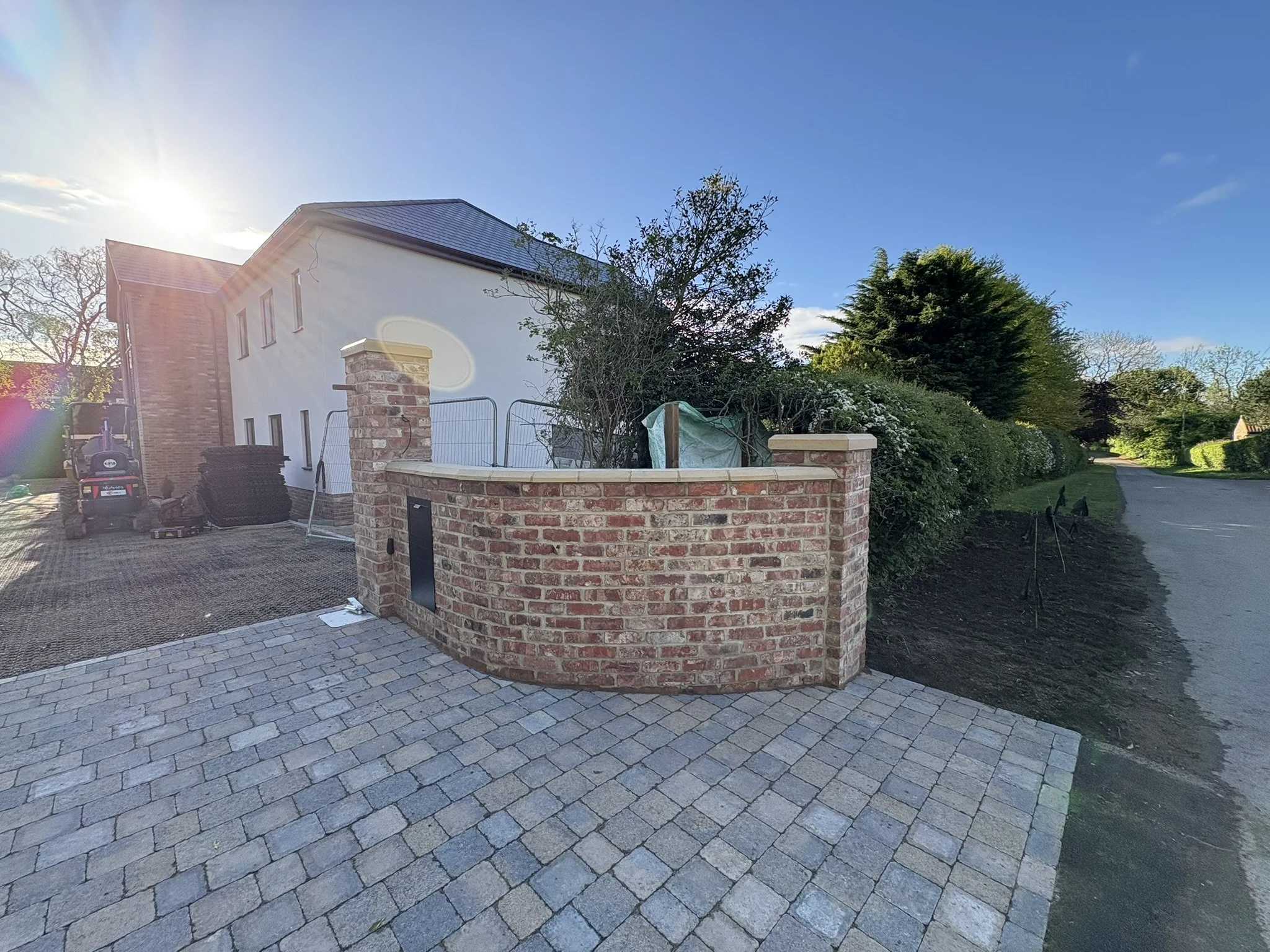 A residential backyard with a paved stone patio, a white house with a metal roof, a brick wall with a gate, and lush green trees and bushes under a bright blue sky.