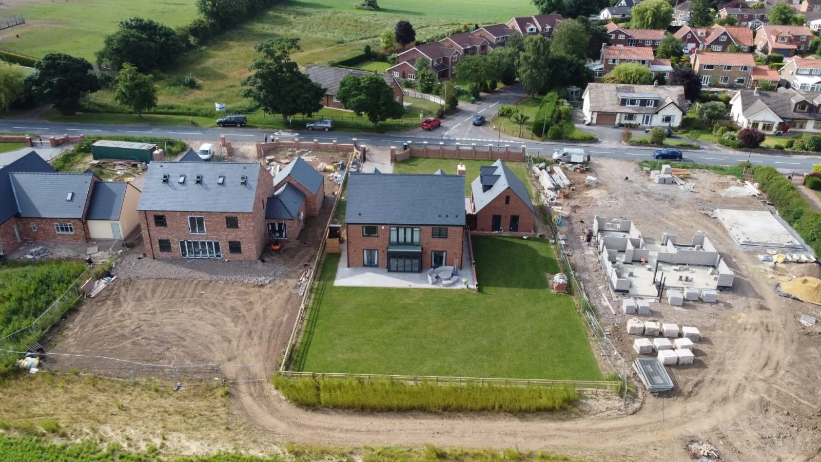 Aerial view of a residential construction site with multiple houses in progress, surrounded by green yards and neighboring homes, with construction materials and vehicles nearby.