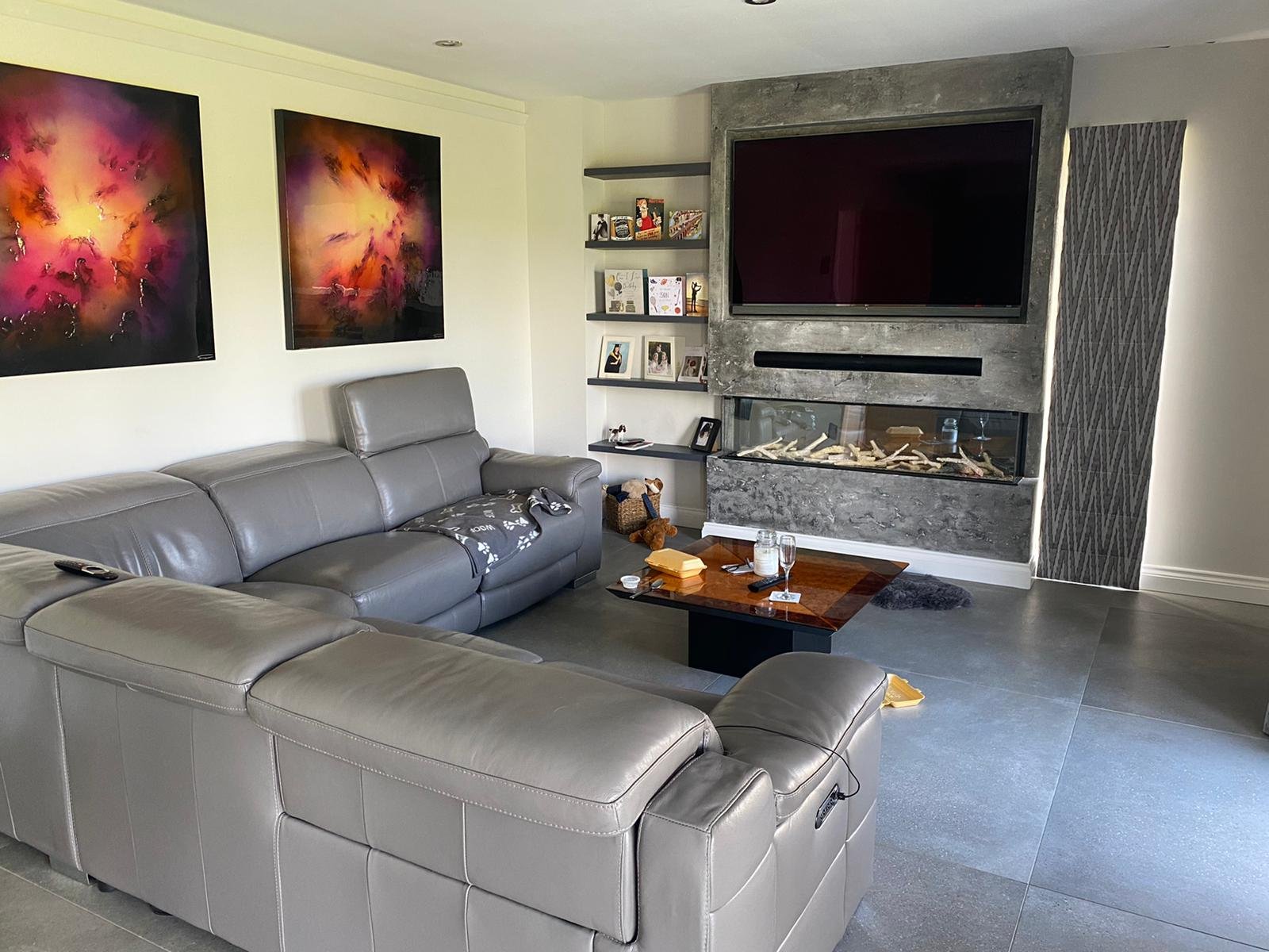Modern living room with grey leather sofas, abstract colorful paintings on the wall, a mounted flat-screen TV, and a fireplace with decor stones, shelves with framed photos, and decorative items.