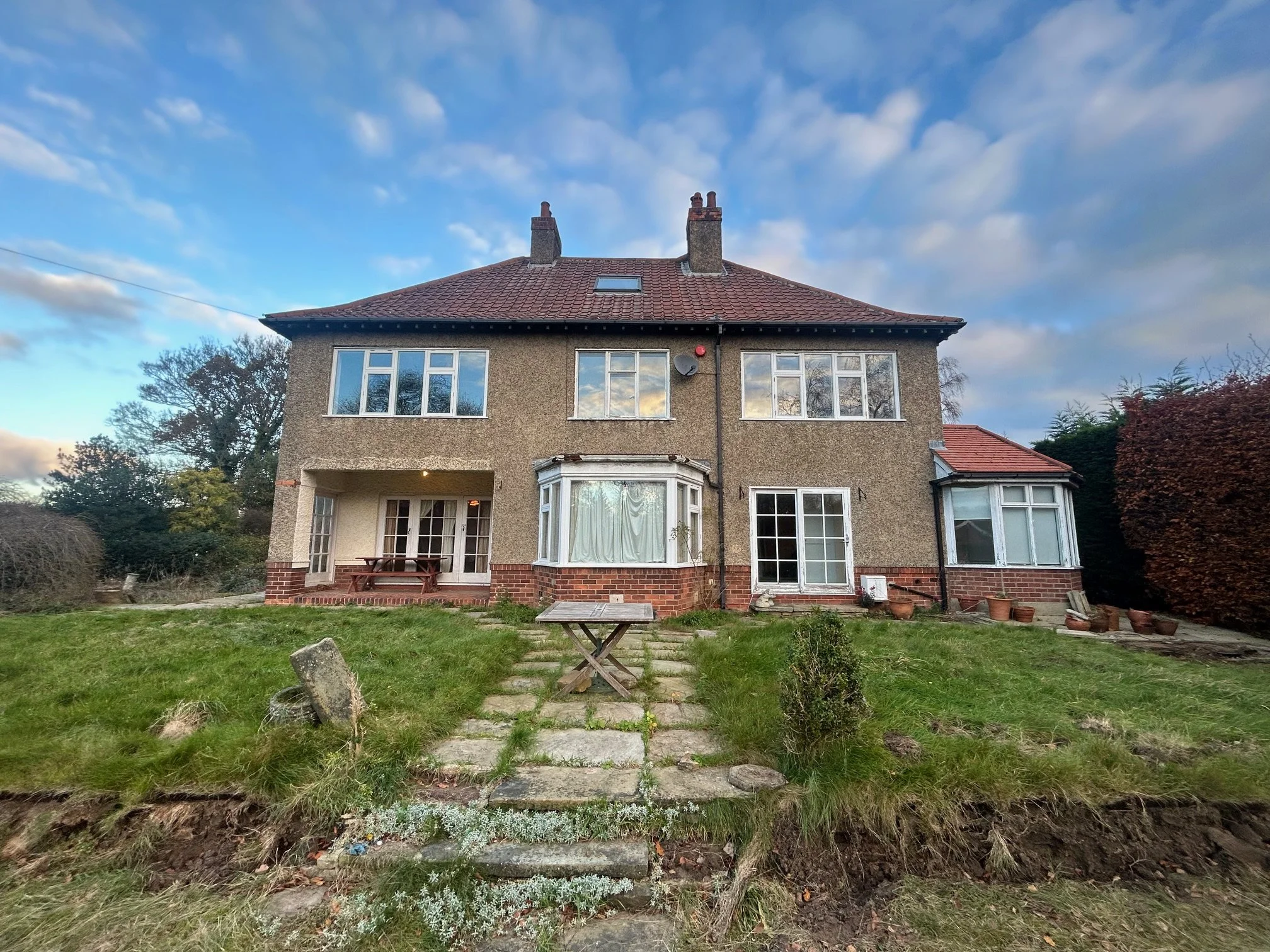 A large two-story house with a brick and stone exterior, multiple windows, and a red-tiled roof, set against a partly cloudy sky.