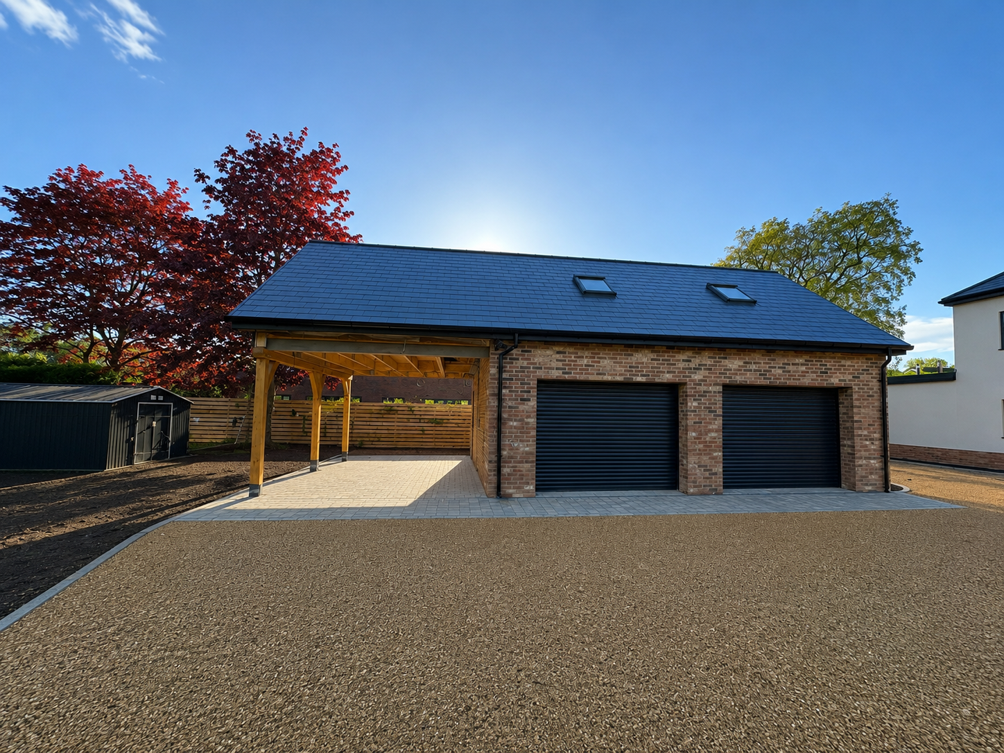Modern detached garage with brick walls, two black roller doors, a wooden covered carport, and a paved driveway, surrounded by trees and a clear blue sky.