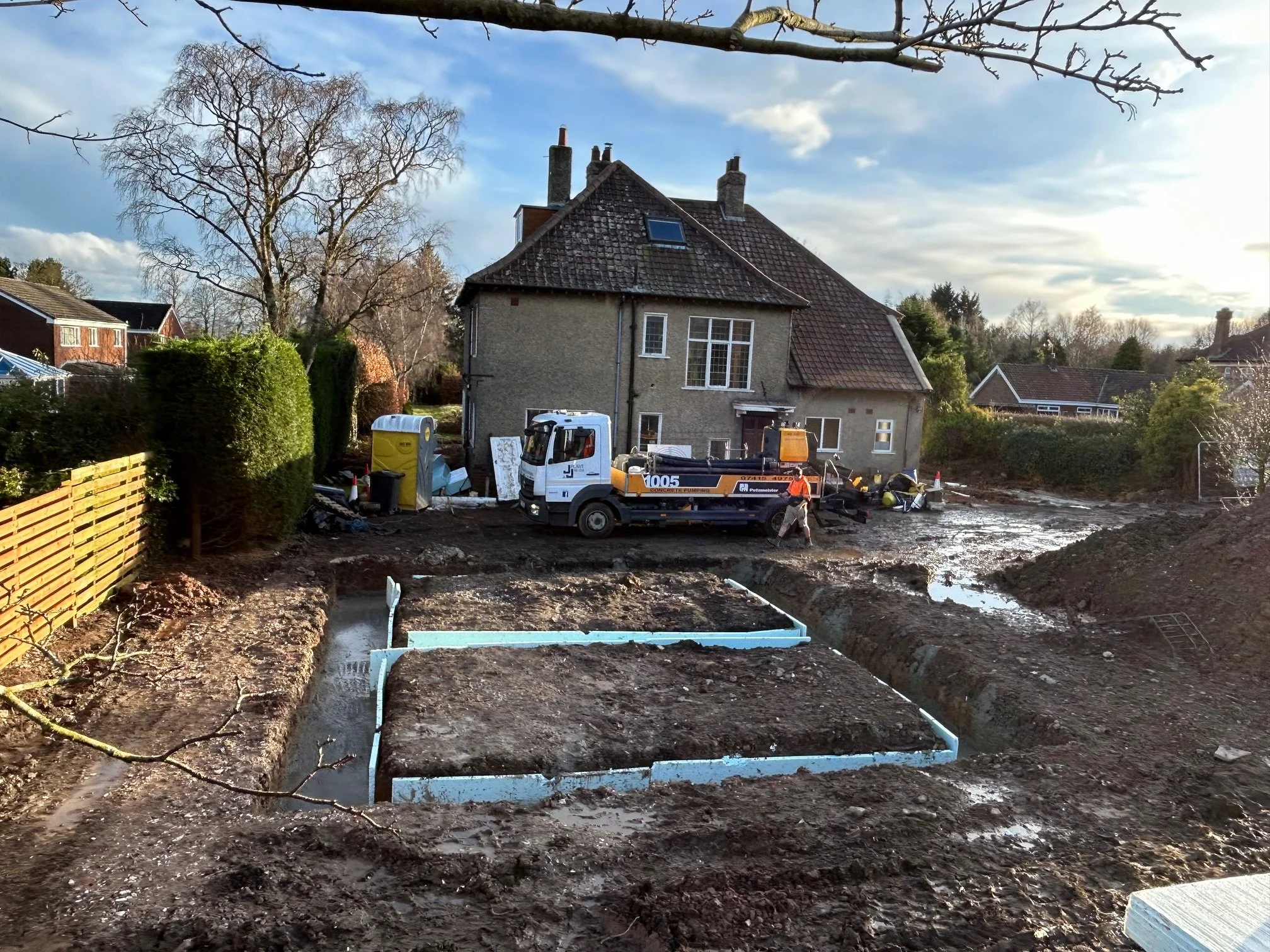 Construction site in front of a house with construction workers, a truck, a portable toilet, and exposed foundation trenches.
