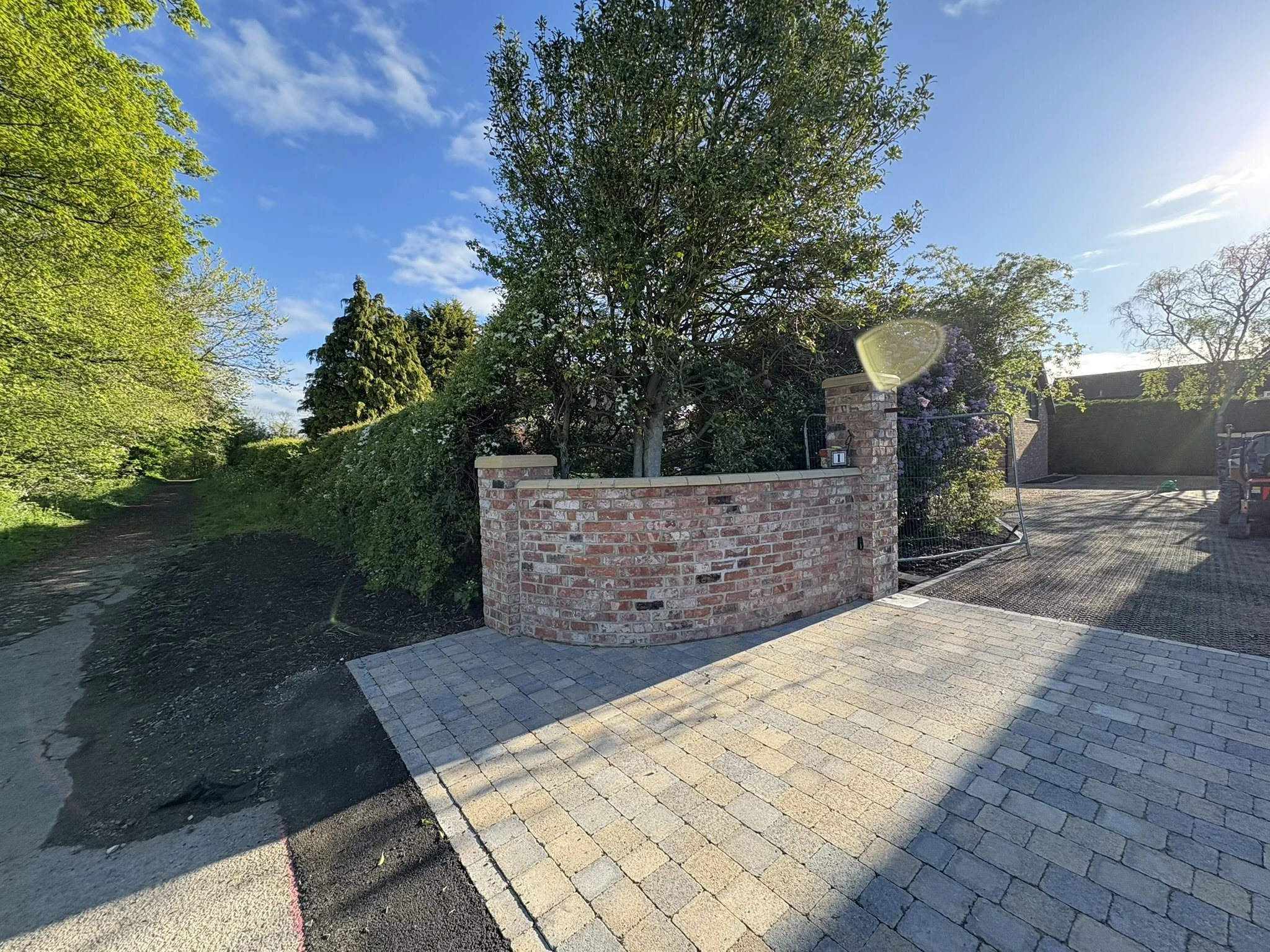 A paved driveway with parking spaces marked, leading to a tall brick wall with a gate, surrounded by greenery and trees in a suburban setting.