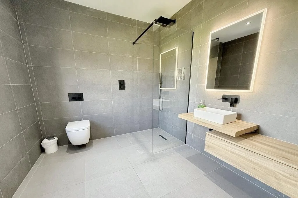 Modern bathroom with gray tiled walls and floor, a wall-mounted toilet, a small waste bin, a walk-in shower with a black rain shower head and a glass partition, a framed mirror with backlighting, a floating wooden vanity with a white rectangular vess