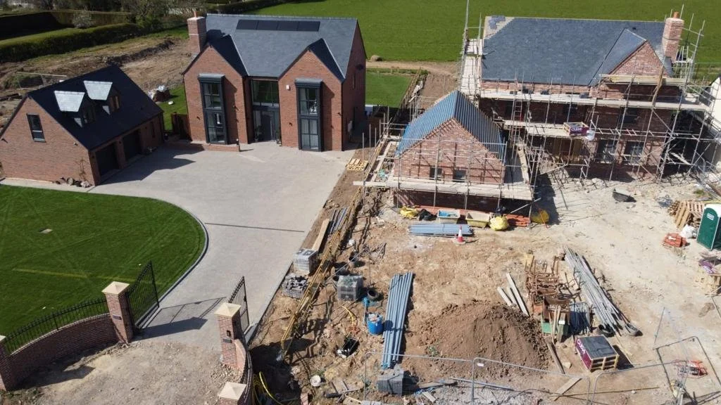 Construction site showing multiple houses under construction, scaffolding, building materials, and cleared land, with one completed brick house in the background.