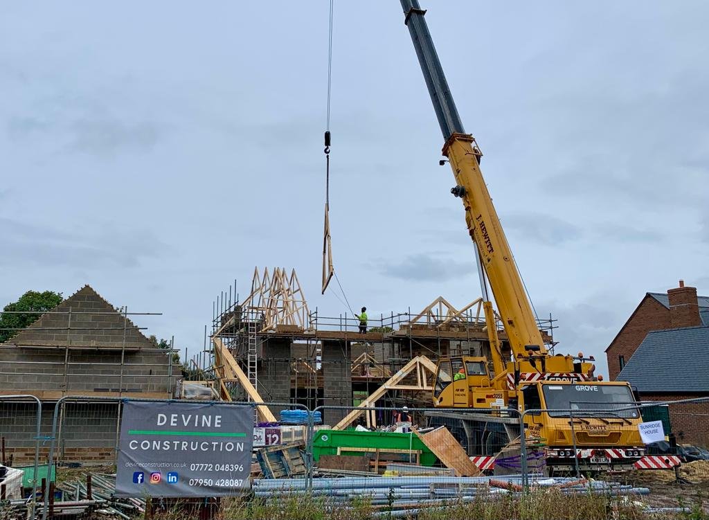 A construction site shows a house being built with a crane lifting wooden beams. Workers are present on the scaffolding and a sign advertisement of Devine Construction is displayed in the foreground.