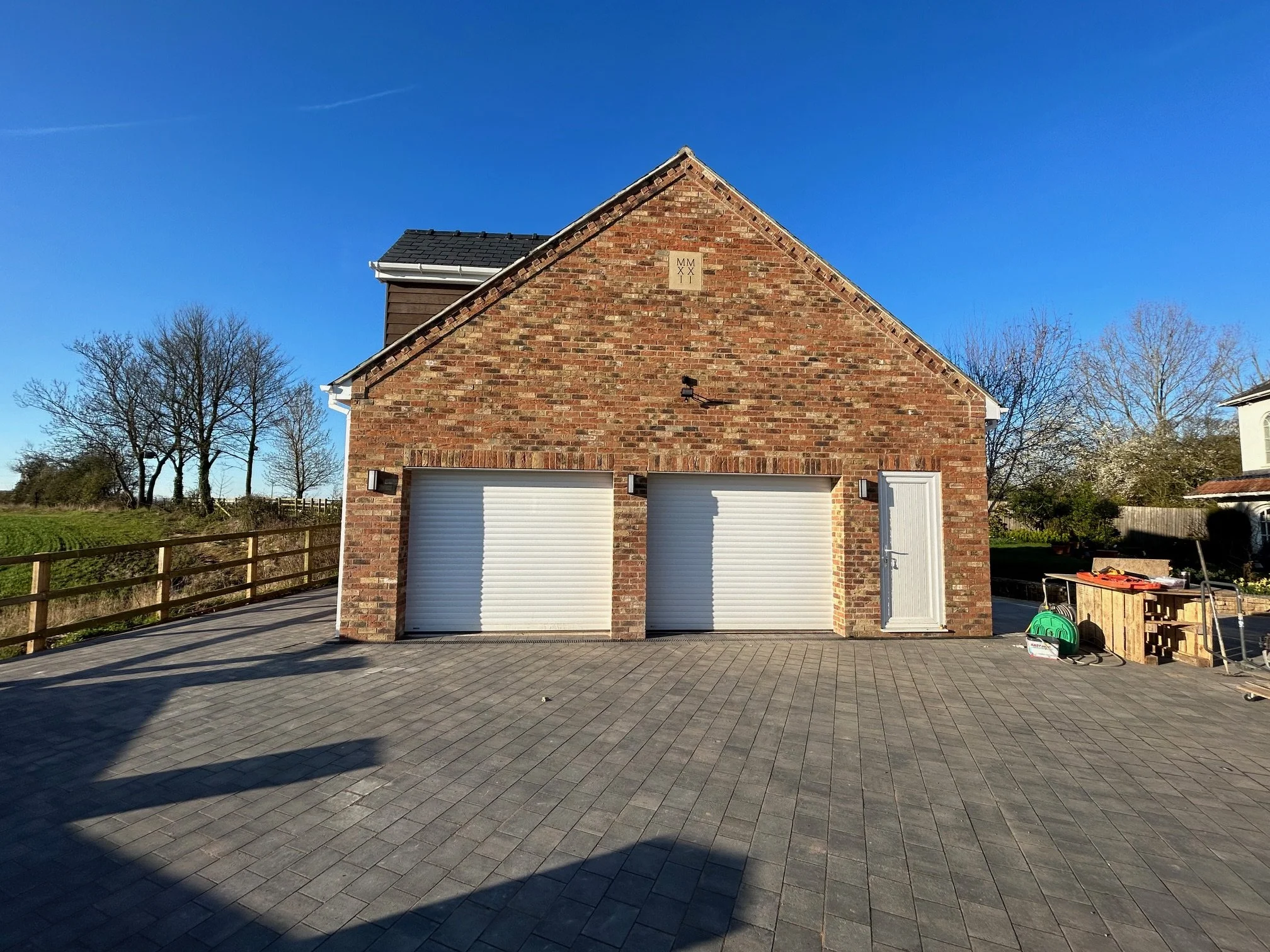 A brick garage with two closed white roll-up doors, a side door, outdoor lights, and a paved driveway under a clear blue sky, with leafless trees in the background.