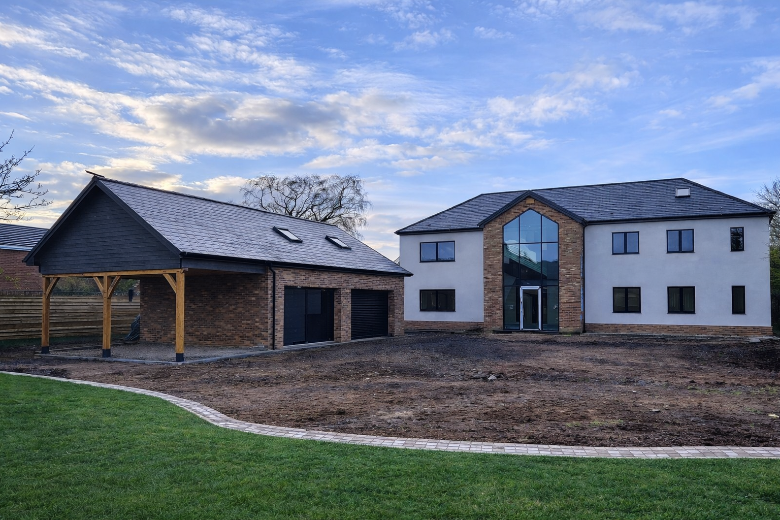 Newly constructed modern house with large glass window and a brick exterior, next to a smaller building with a sloped roof, in a yard with green grass and a paved walking path, under a partly cloudy sky.