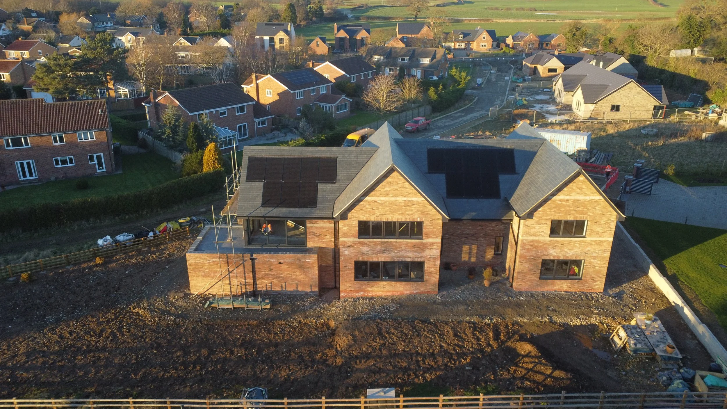 An aerial view of a residential area showing a house under construction with brick walls and solar panels on the roof, surrounded by other completed homes, trees, and green fields.