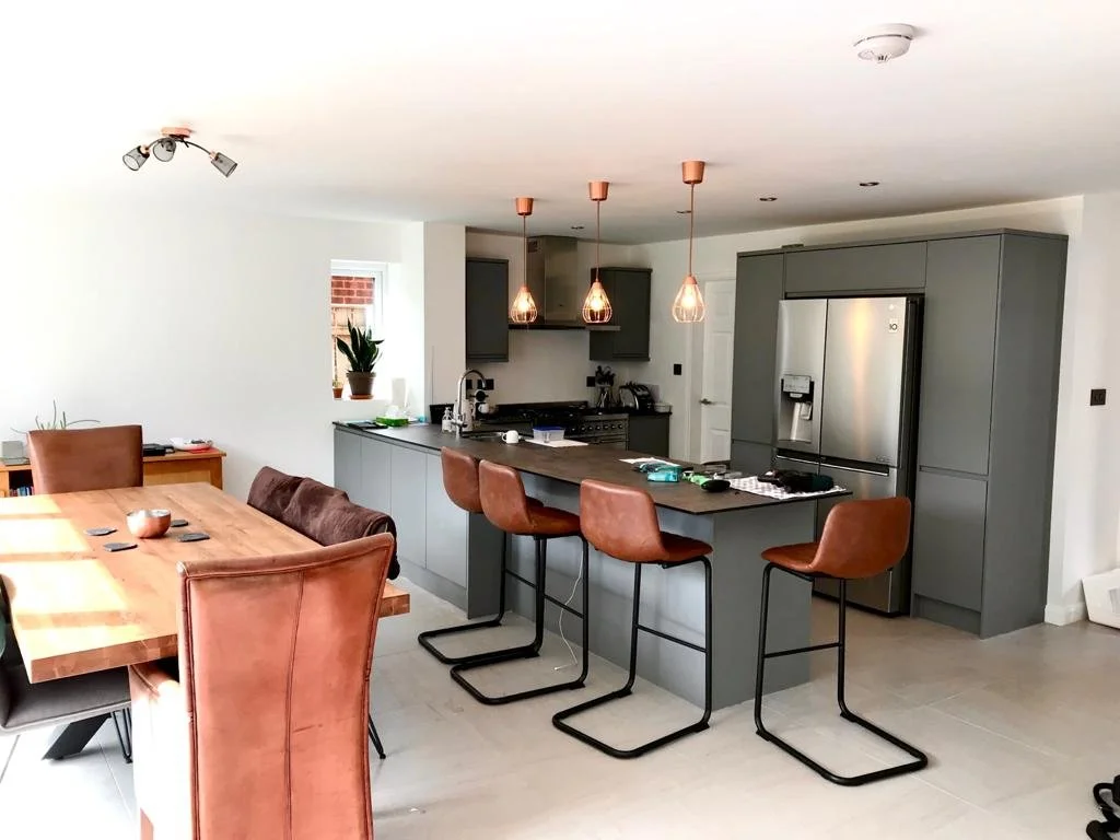 Modern kitchen and dining area with a large wooden table, brown dining chairs, and a kitchen island with three brown barstools. The kitchen has gray cabinets, a stainless steel refrigerator, and pendant lighting.
