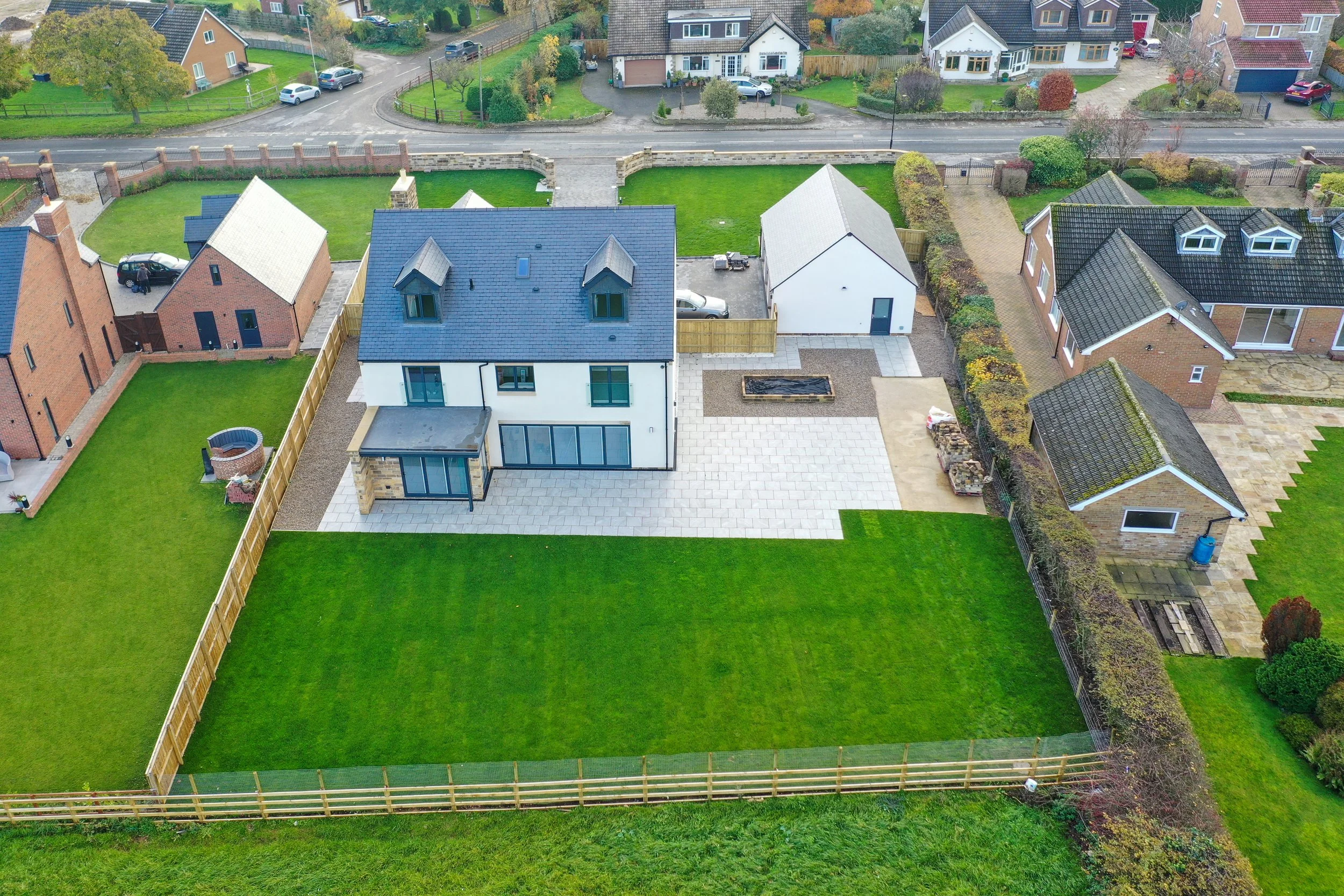An aerial view of a modern two-story house with a blue roof, white walls, large windows, and a backyard with a green lawn, paved patio, and a small garden bed, in a suburban neighborhood.