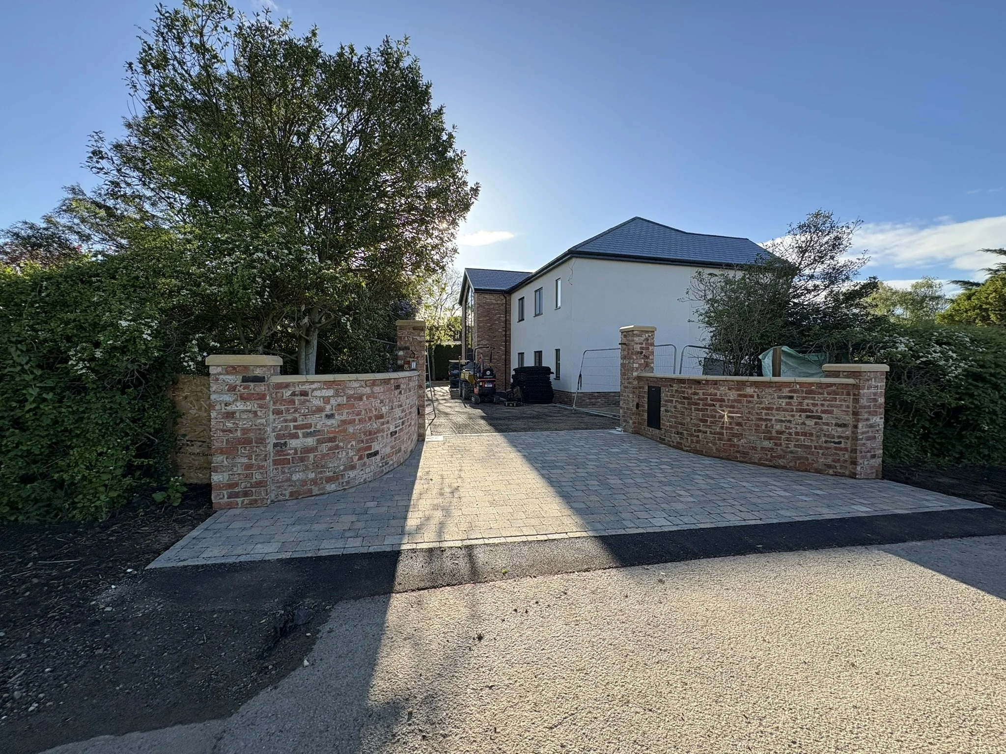 A newly paved driveway with a brick wall on both sides, leading to a modern two-story house with white exterior walls, dark roof, and several windows. There are trees and construction materials or equipment on the driveway.
