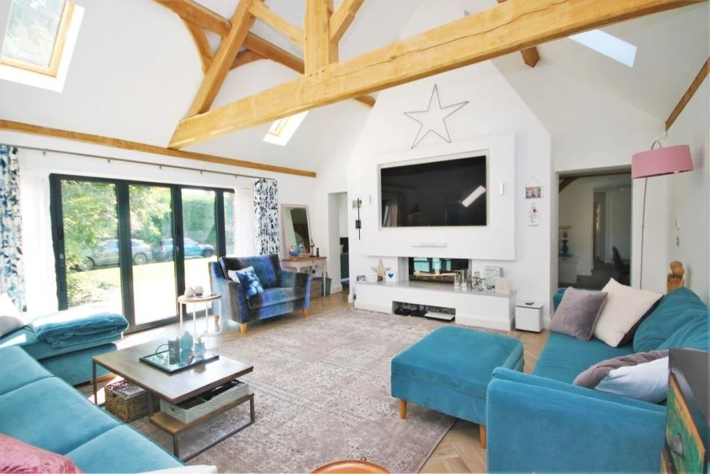 Living room with teal and blue sofas, a wooden coffee table, large window with floral curtains, a fireplace, mounted flat-screen TV, and exposed wooden beams on the ceiling.