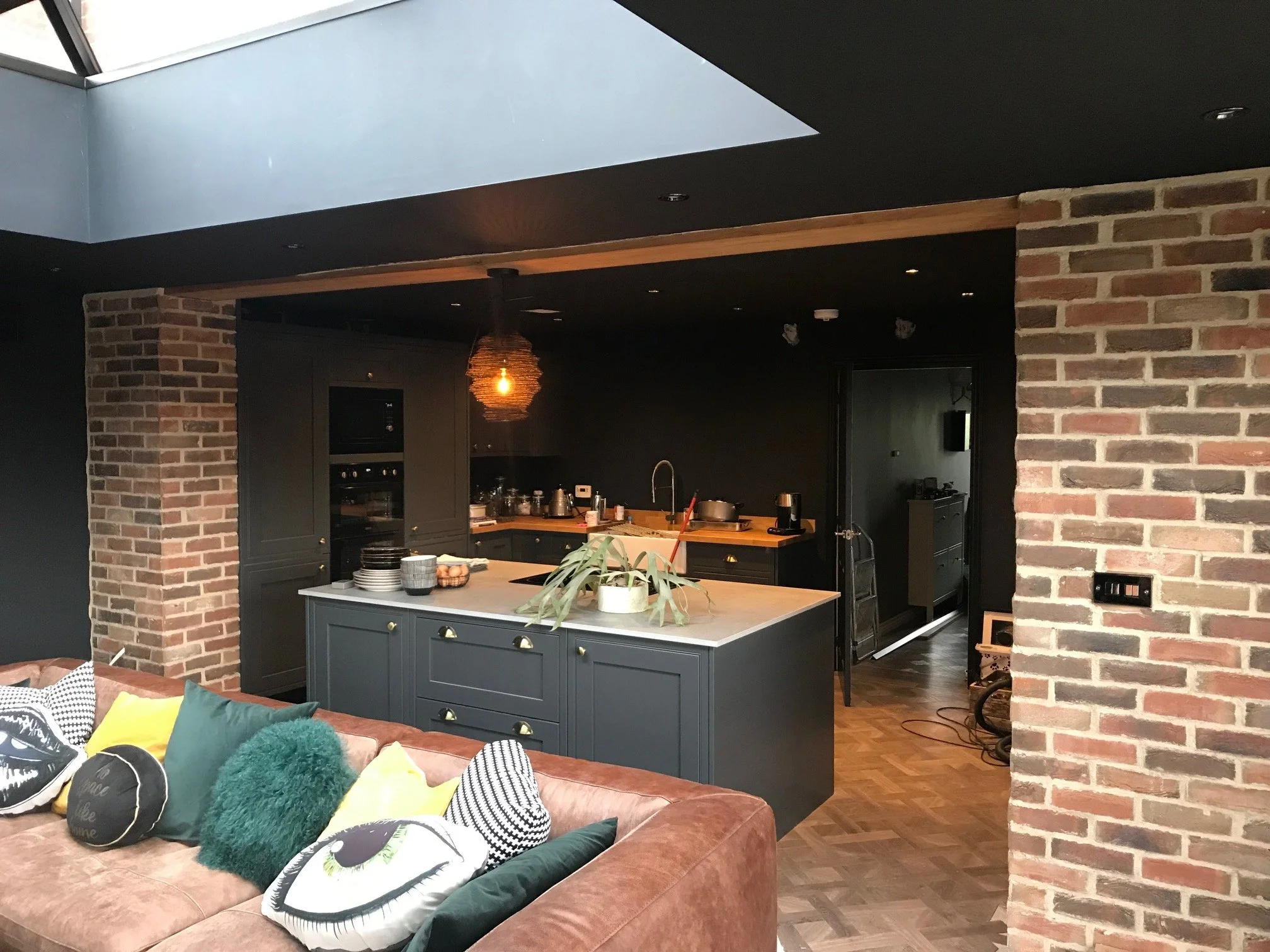 Open-concept living room and kitchen area with exposed brick walls, dark cabinetry, a large island with a light-colored countertop, assorted decor, and a sofa with decorative pillows.