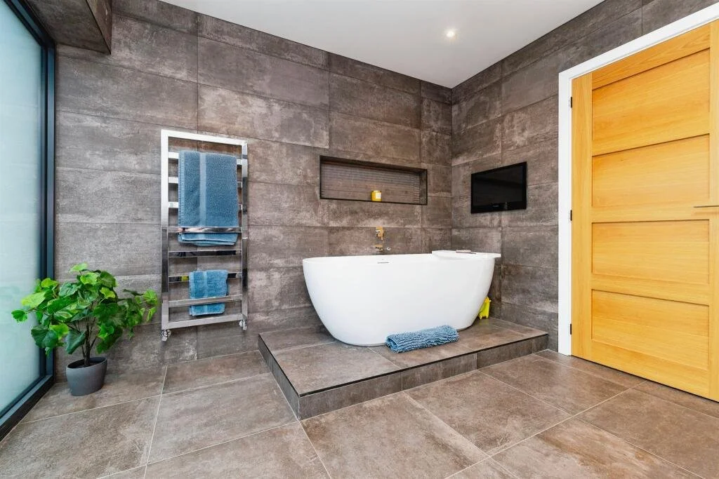 Modern bathroom with gray tiled walls and floor, a white freestanding bathtub on a slightly elevated platform, a small wall-mounted TV, a towel rack with blue towels, a potted plant, a small recessed shelf with a candle, and a wooden door.