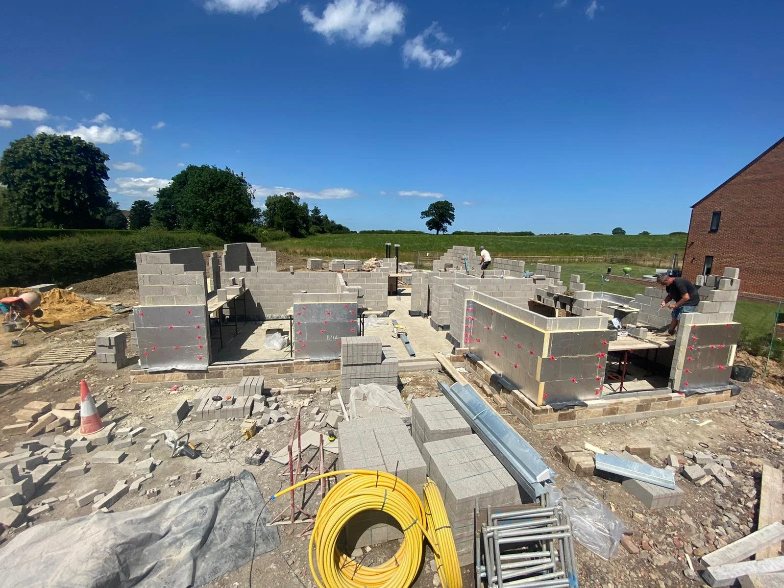 Construction workers building the foundation of a house with gray concrete blocks, outdoor setting with trees and open sky.
