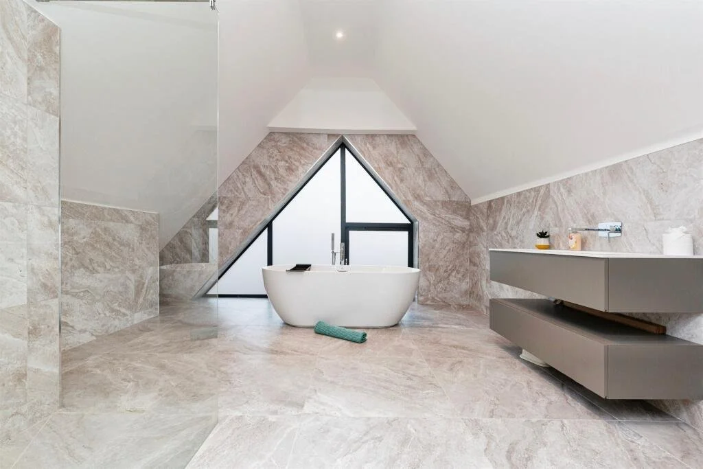 Modern bathroom with a freestanding bathtub, triangular window, and beige marble walls and floors.