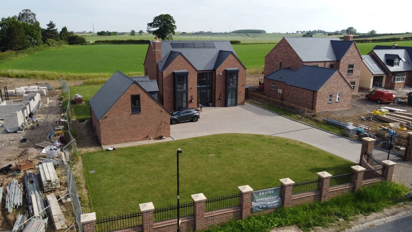 A new brick house under construction with a paved driveway, surrounded by a low brick fence and a green lawn, located in a rural area with fields and trees in the background.
