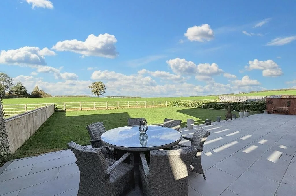 A spacious outdoor patio with a round glass table and six wicker chairs, overlooking a green lawn with a fence and open fields under a bright blue sky with scattered clouds.