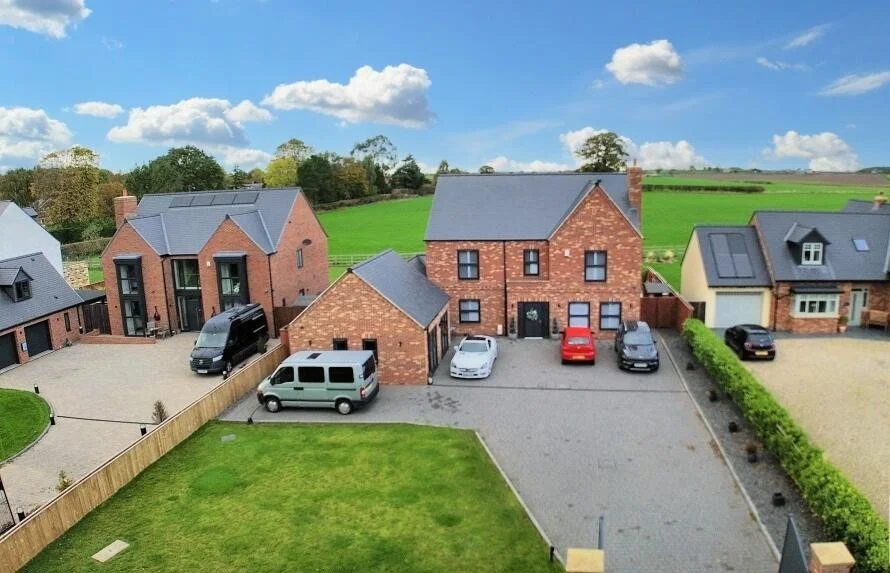 A row of brick houses with modern architecture in a suburban neighborhood, with a fenced lawn in the front and parked cars in the driveway and street. Green fields and trees are in the background under a partly cloudy sky.