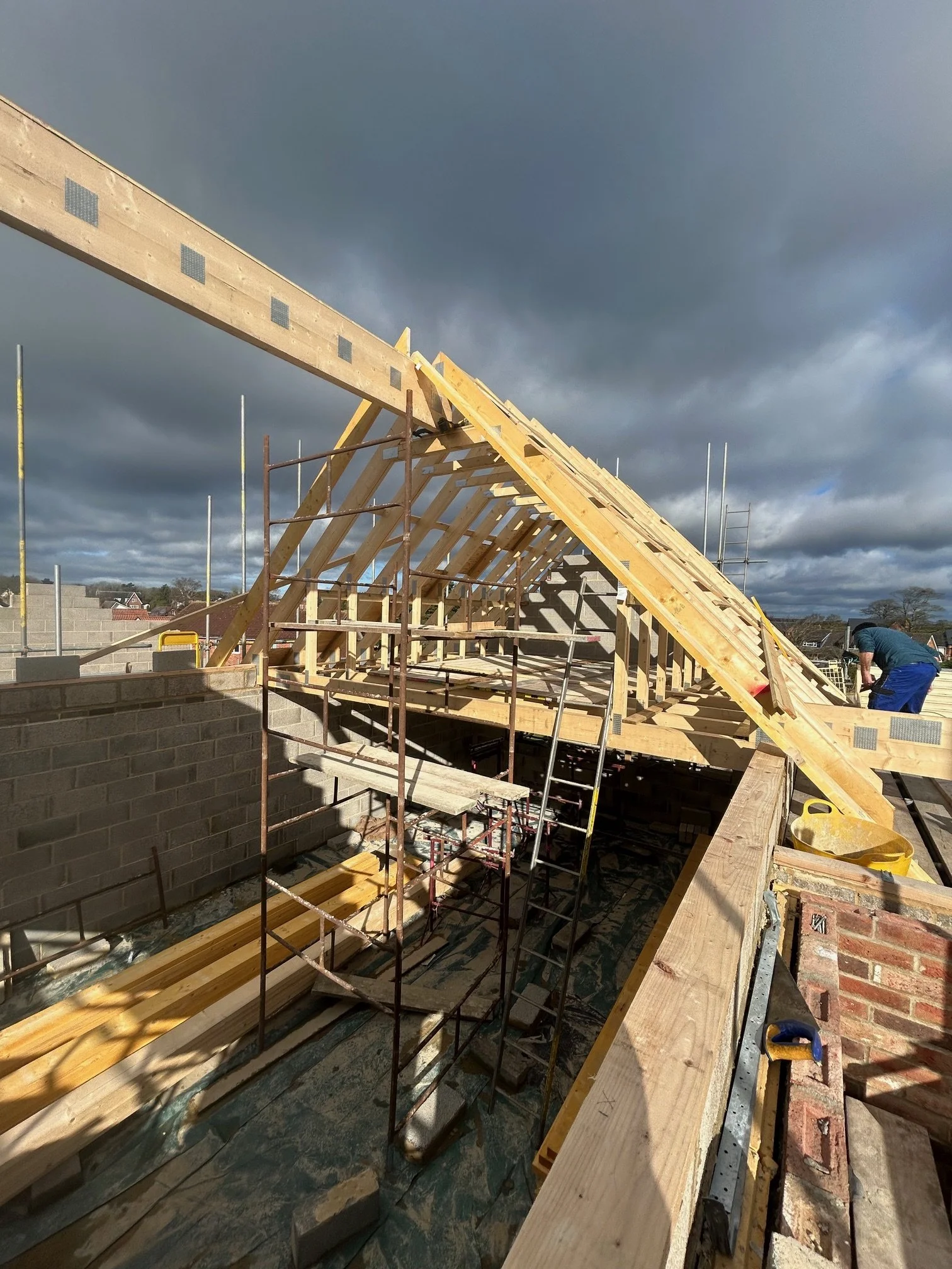 Construction site with wooden roof framework and scaffolding, worker on the right, dark cloudy sky overhead.