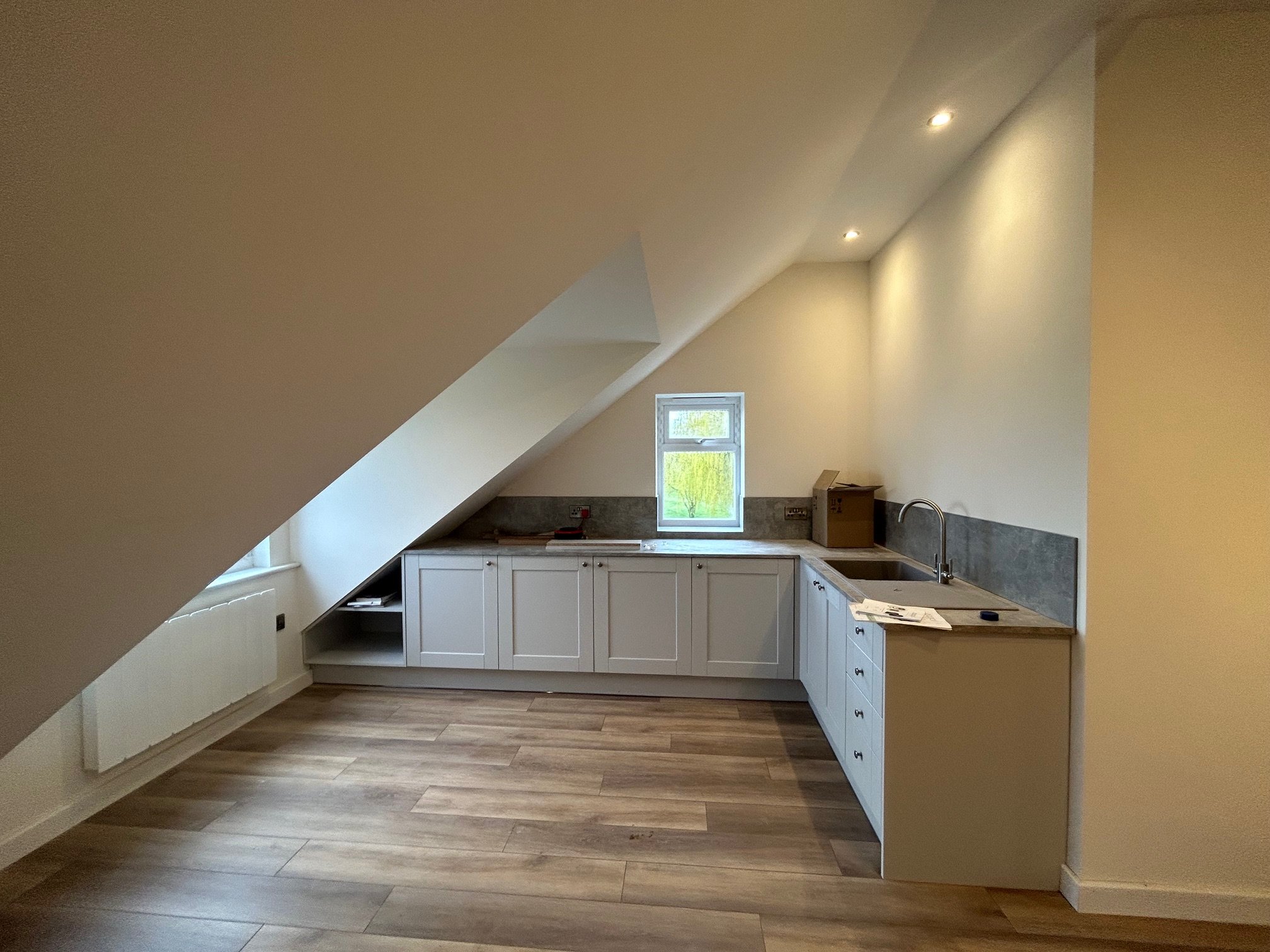 An unfinished attic kitchen with white cabinets, a small window, and a sink, under a sloped ceiling with recessed lighting.