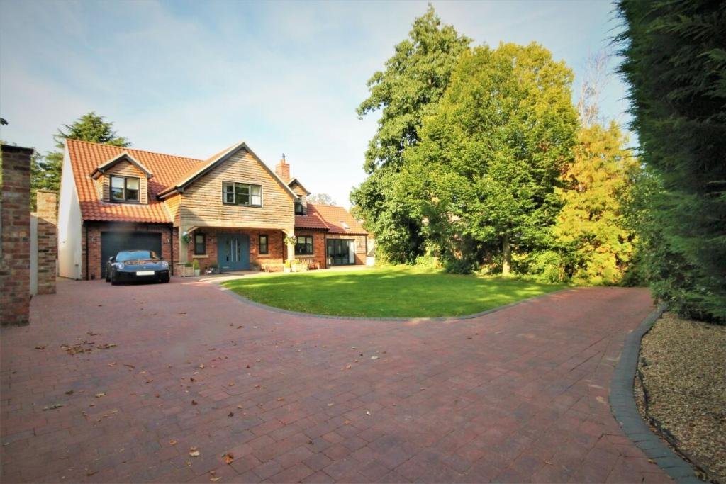 A large house with a red tiled roof, brick and wooden exterior, and blue front door, situated in a green yard with a curved red brick driveway and big trees in the background.