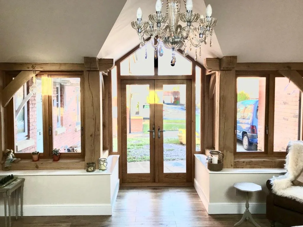Interior view of a room with a wooden front door, large windows, a chandelier, and a small side table with decorative items.