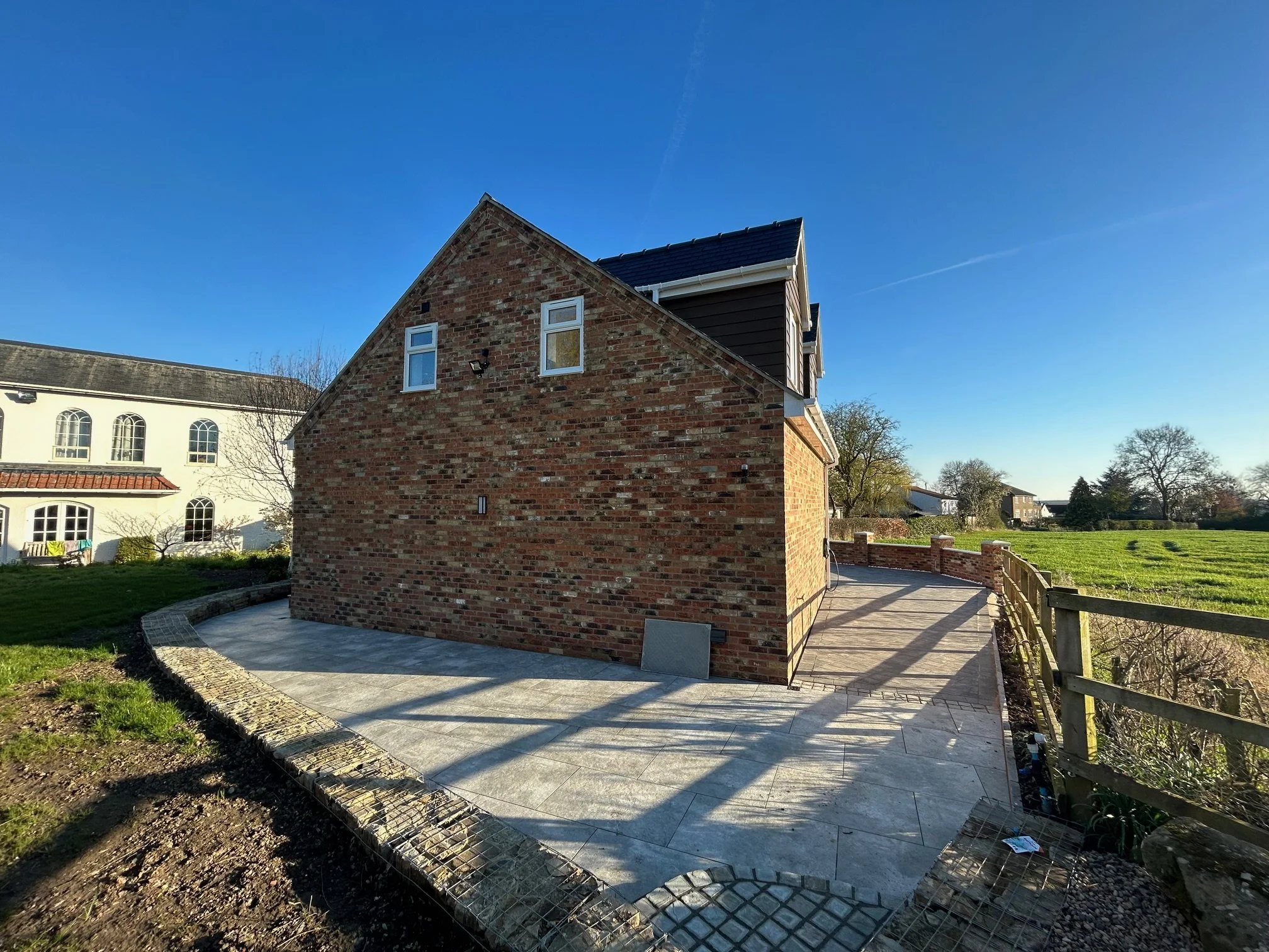 Backyard with newly constructed brick house, newly paved concrete patio, wooden fence, green grass lawn, clear blue sky, trees in the background.