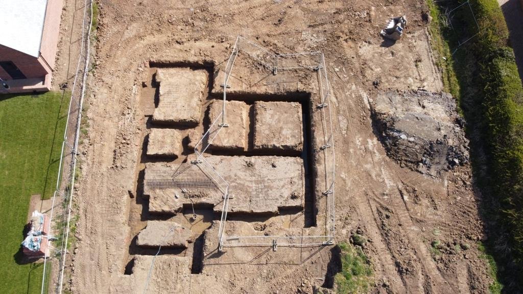 Aerial view of an archaeological excavation site with exposed ancient stone foundation walls and trenches, surrounded by dirt and construction equipment.