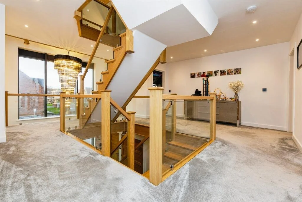 Living area with beige carpet, staircase with wooden railing and glass panels, large window, modern chandelier, and a desk with decorative items against the wall.