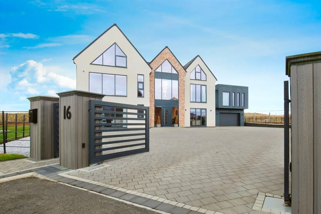 Modern multi-story house with large windows, brick accents, and a gated driveway with the number 16, set against a clear blue sky.