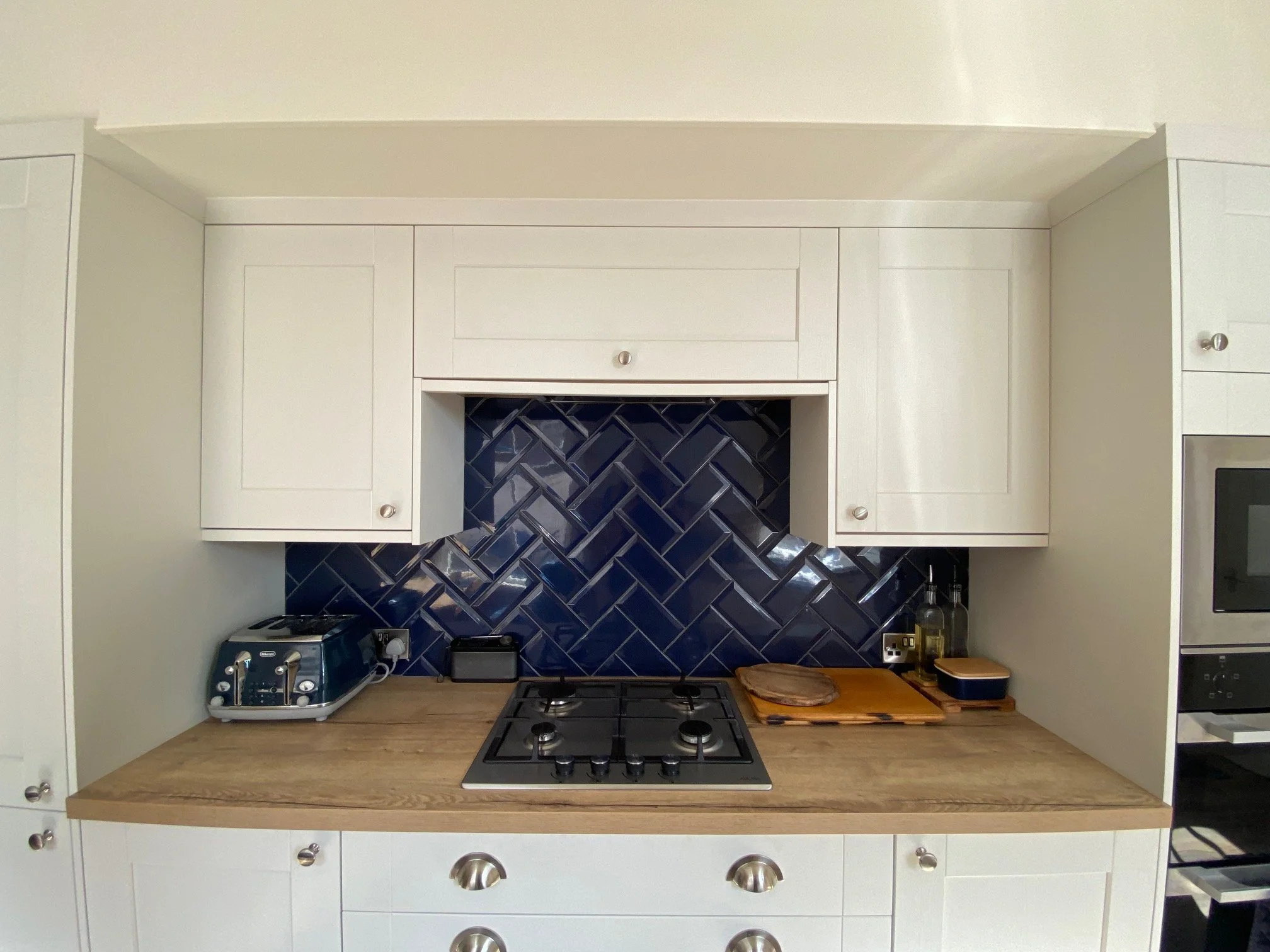 Kitchen with white cabinets, wood countertop, blue herringbone tile backsplash, gas stove, toaster, cutting board with bread, and bottles on the counter.