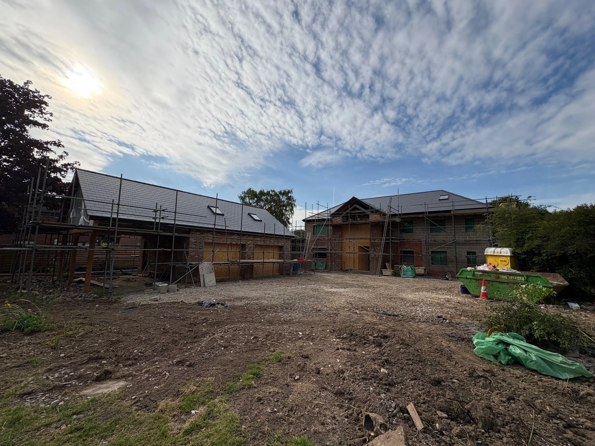 Construction site with two brick buildings under construction, scaffolding around them, and a dirt ground in the foreground. The sky is partly cloudy with the sun partially visible.