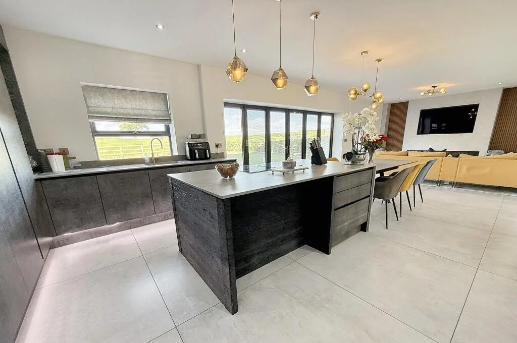Modern open-concept kitchen and living area with black island, gray cabinets, pendant lights, dining table with chairs, and a large TV on the wall.