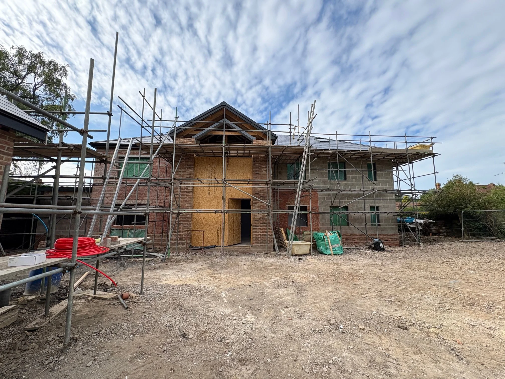 A house under construction with scaffolding around it, some parts covered with plywood, and construction materials on the ground.