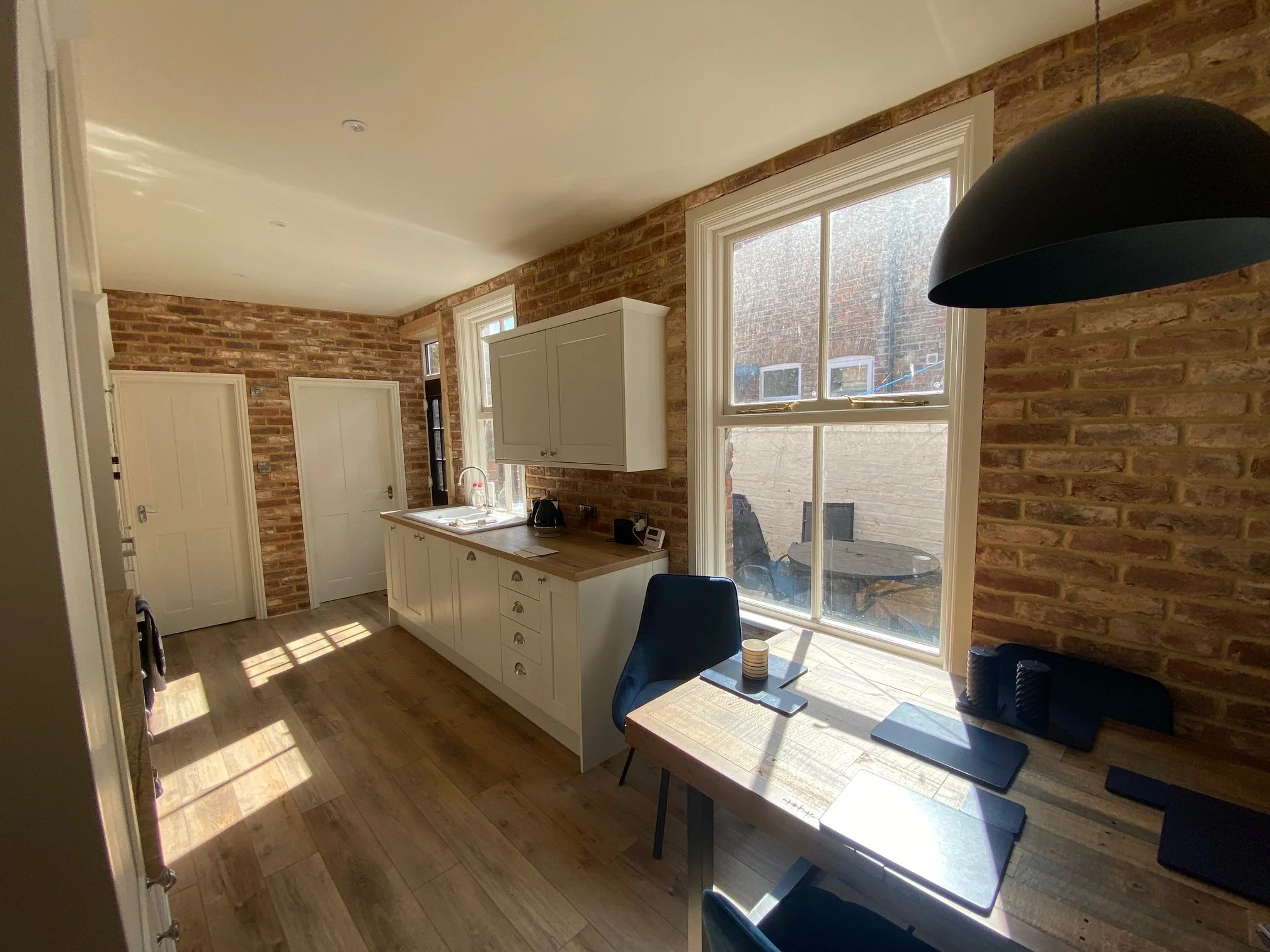 Kitchen with brick walls, white cabinets, wooden countertop, black chairs, large window letting in sunlight, and a table with a laptop and candles, overlooking a small outdoor patio with a table and chairs.