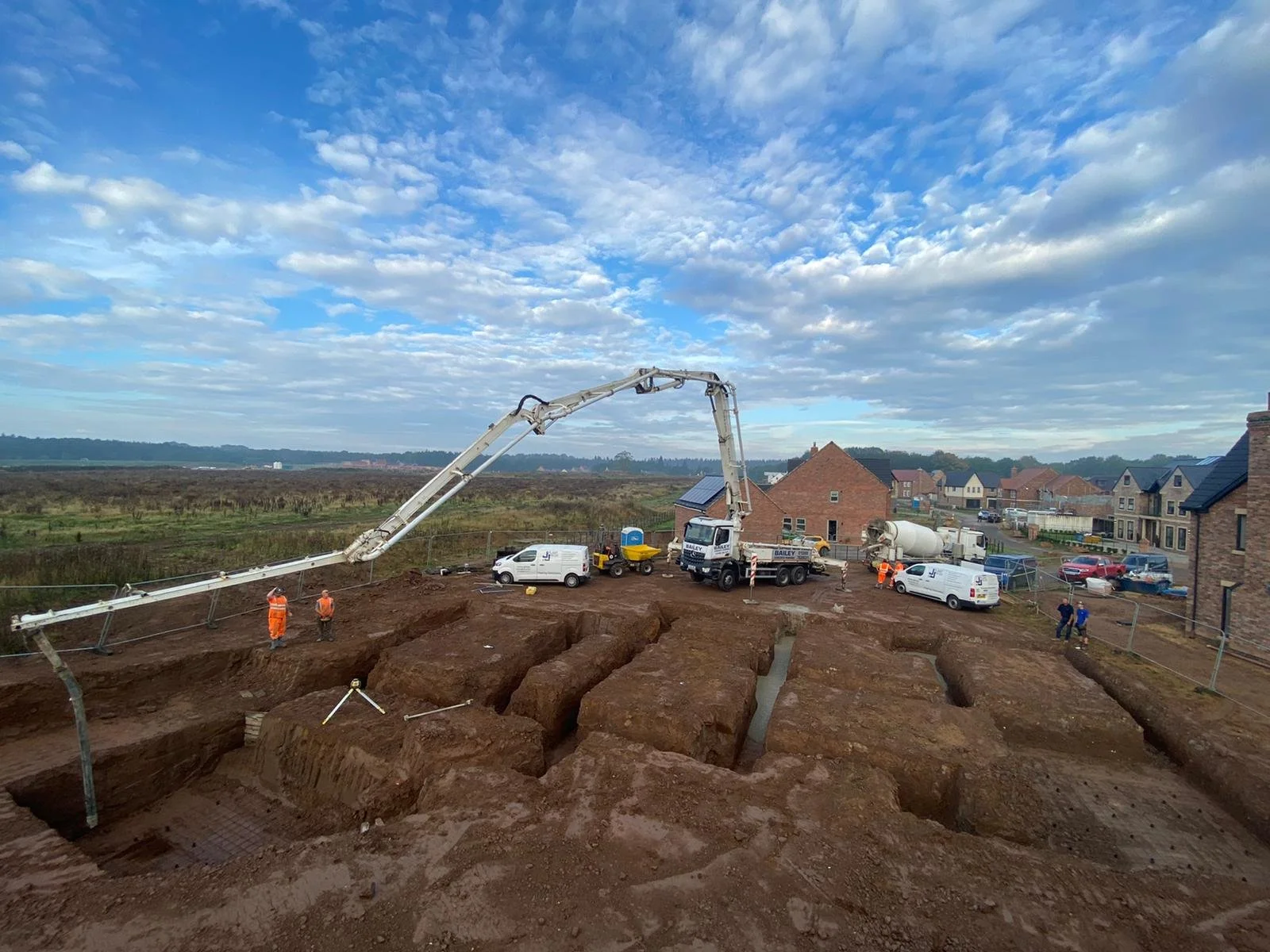 Construction site with trucks and workers digging trenches in a residential area, with houses and a cloudy sky in the background.