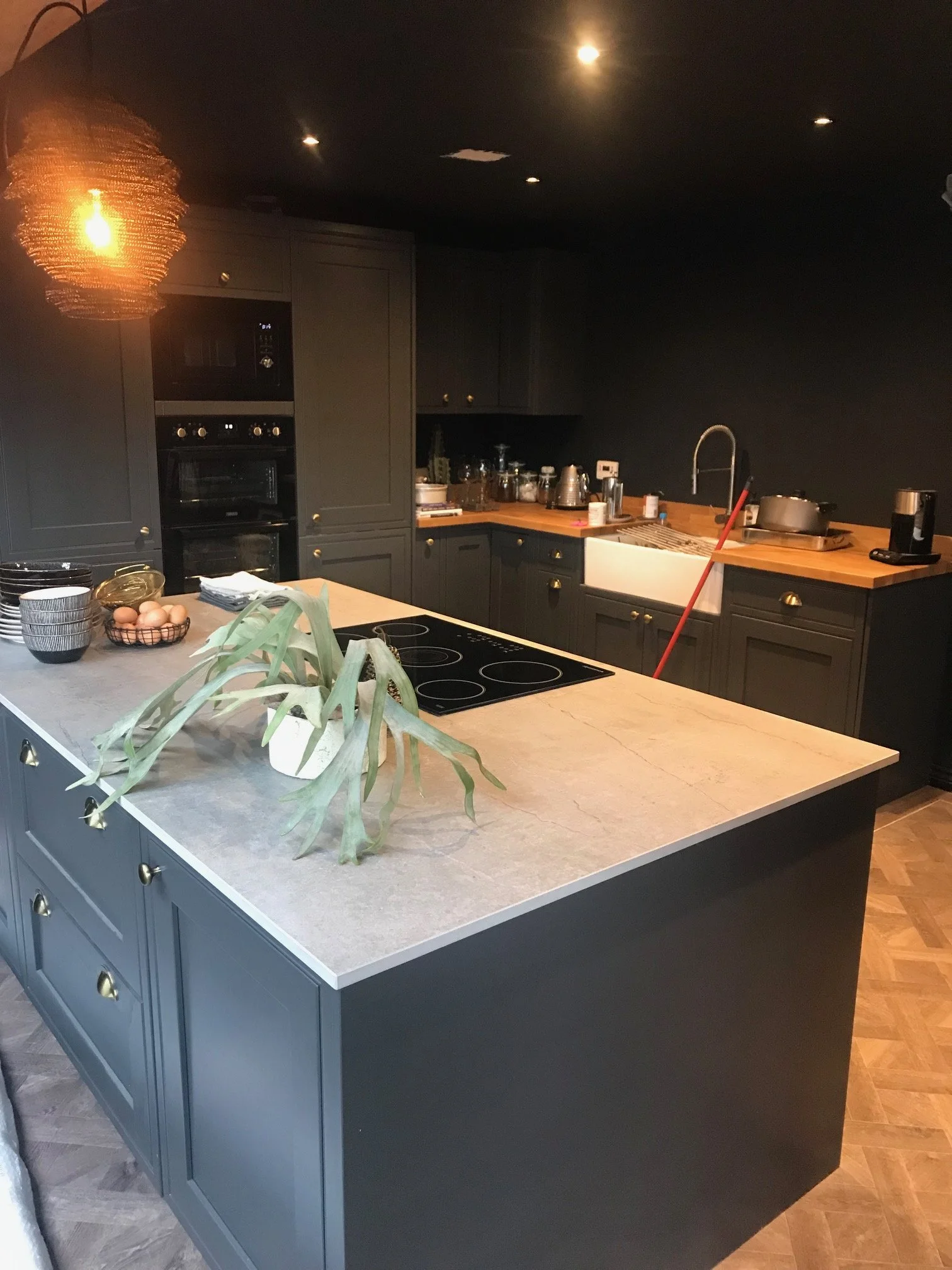 Modern kitchen with dark cabinets, white countertop island, wooden countertop on the back wall, and black walls. The island has a potted plant, bowls of eggs, and napkins. Appliances include a microwave, oven, and stovetop. Kitchen utensils and dishe