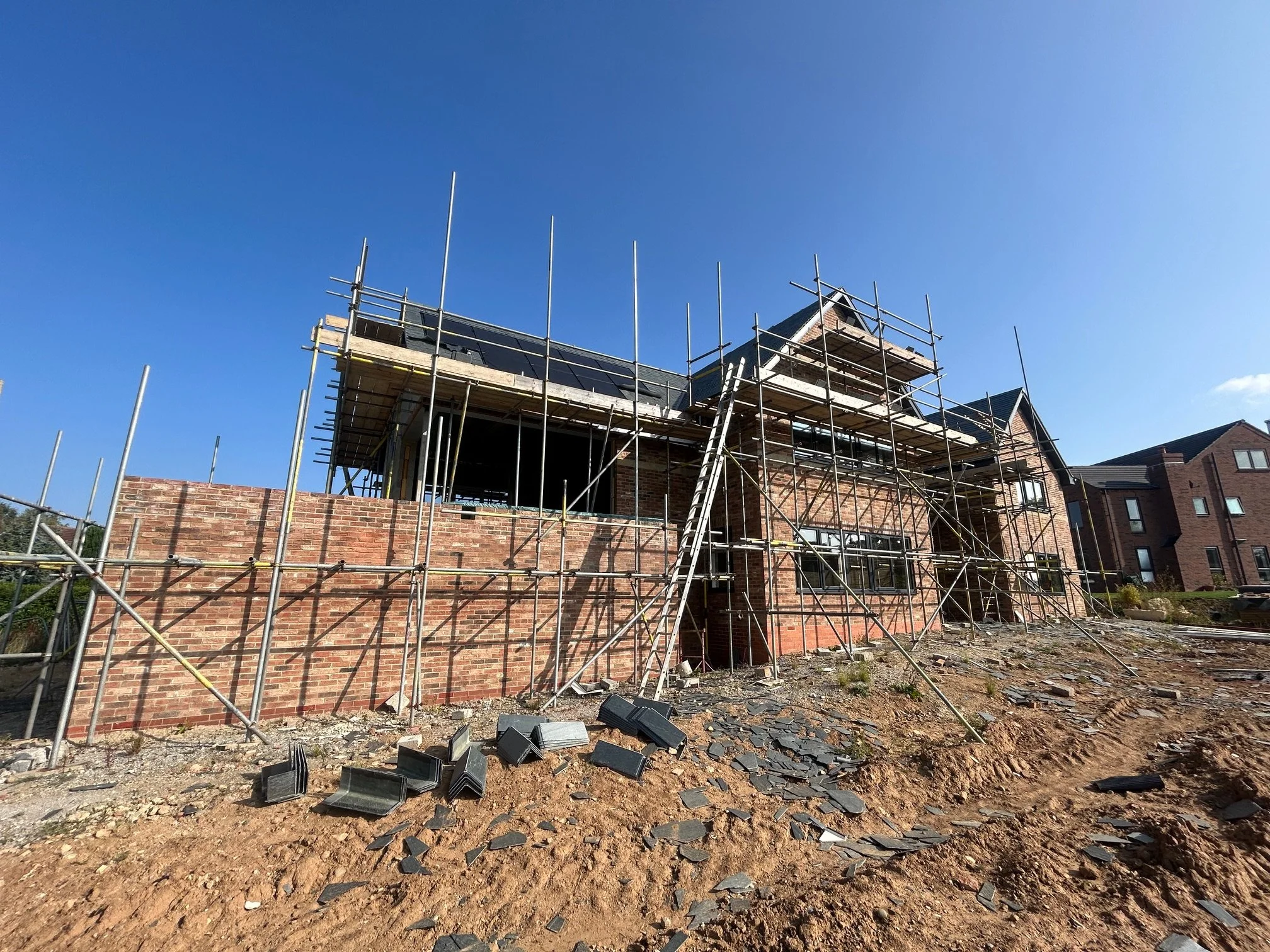 Construction site with scaffolding around a brick house, some roof tiles on the ground, and a clear blue sky.