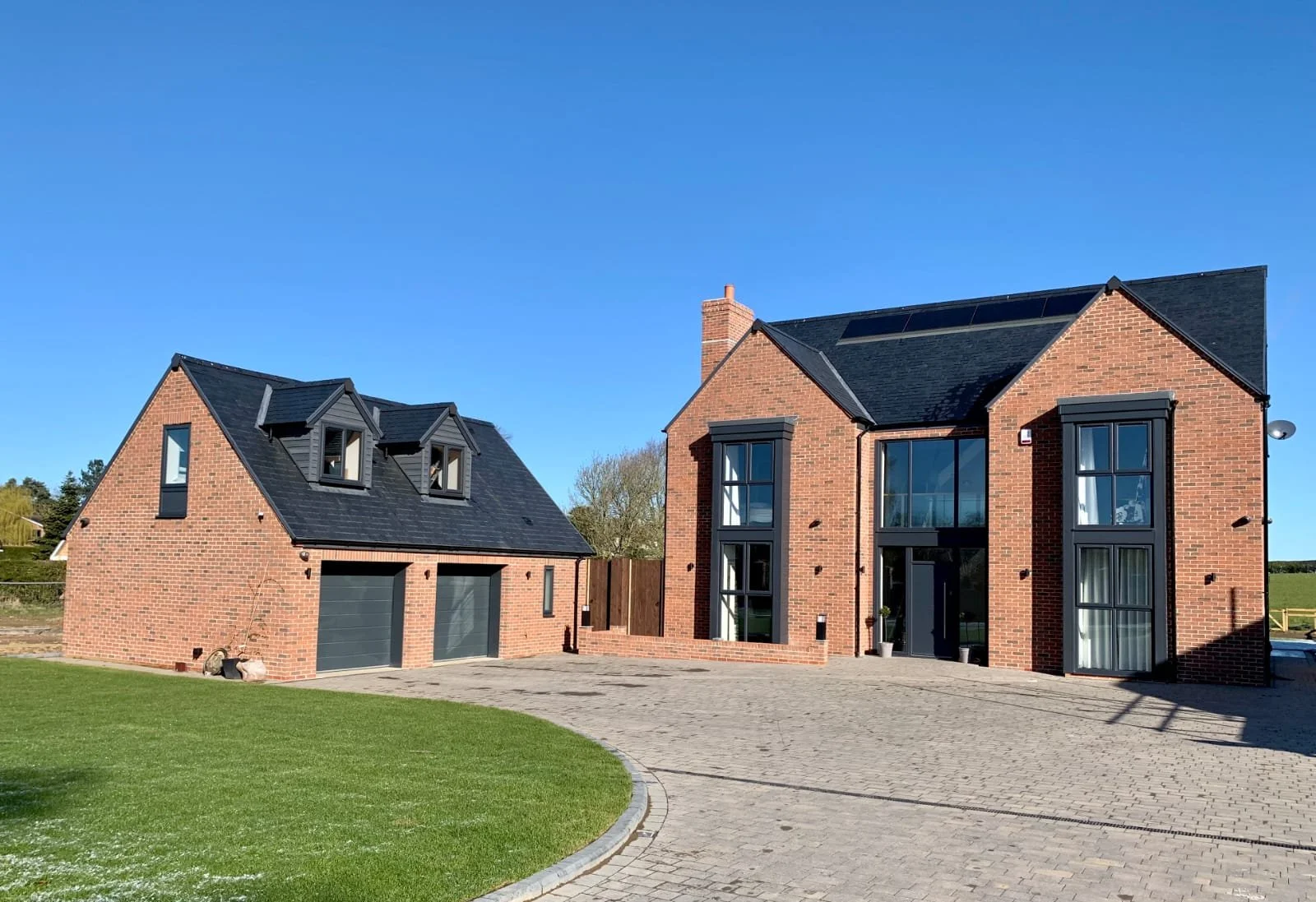 A modern brick house with large windows and a black roof, accompanied by a detached garage with dormer windows, situated on a well-maintained lawn and paved driveway under a clear blue sky.