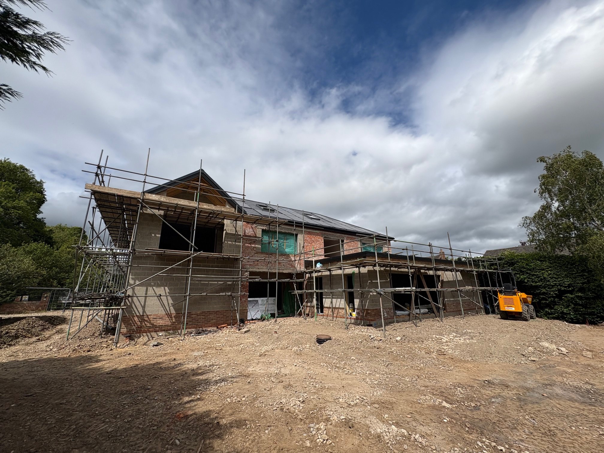 A two-story house under construction with scaffolding around it, a yellow construction vehicle parked to the right, and trees on either side. The sky is partly cloudy.