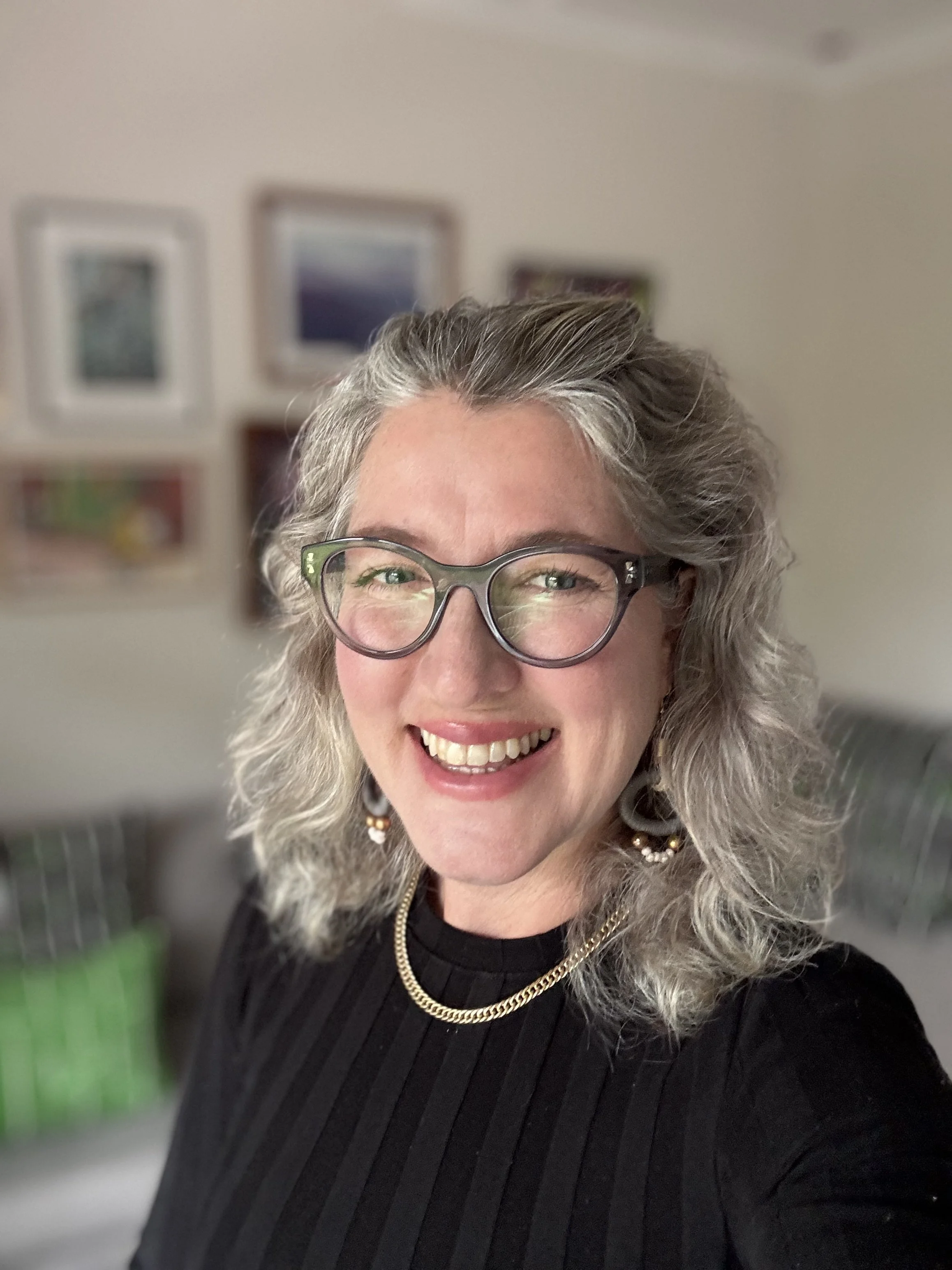 Neurodiversity Coach Suna Hall - A smiling woman with gray curly hair, glasses, earrings, and a gold chain necklace, wearing a black ribbed top, in an indoor setting with framed pictures on the wall in the background.