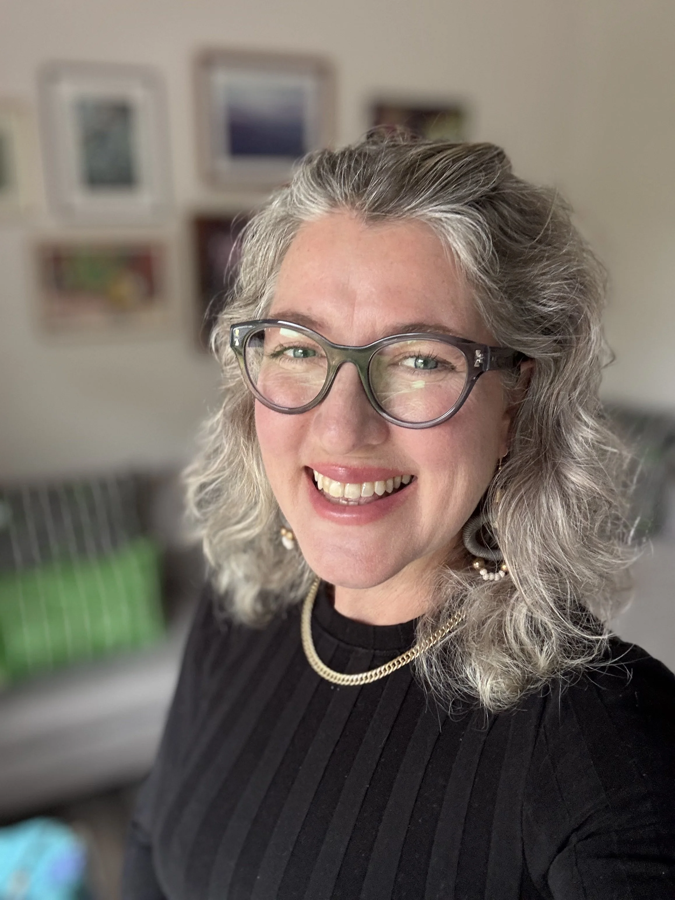 Neurodiversity Coach Suna Hall - A smiling woman with gray curly hair, glasses, earrings, and a gold chain necklace, wearing a black ribbed top, in an indoor setting with framed pictures on the wall in the background.