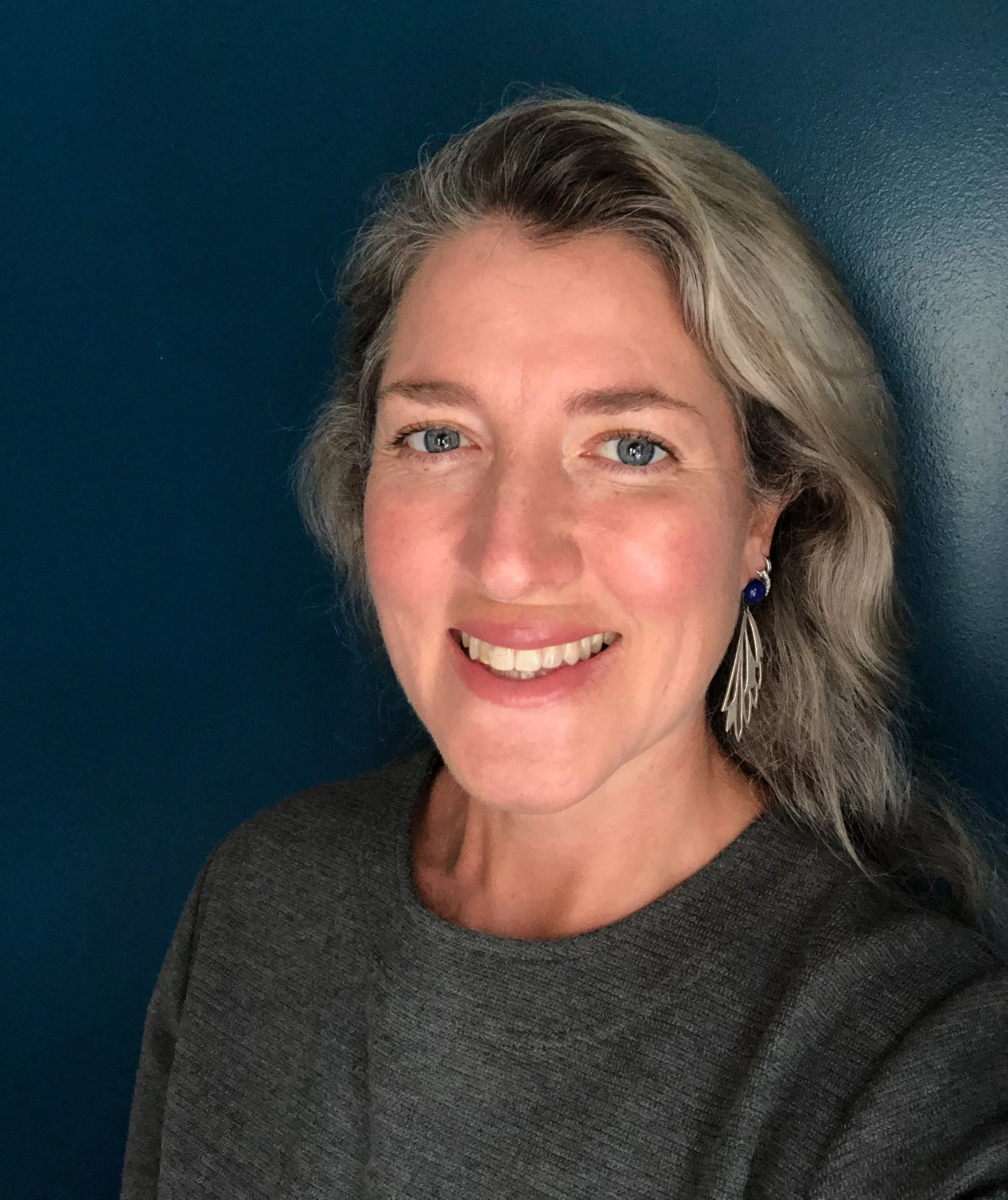 Close-up of Suna Hall, Neurodiversity Coach - a smiling woman with blonde hair, blue eyes, and earrings, against a dark teal background.